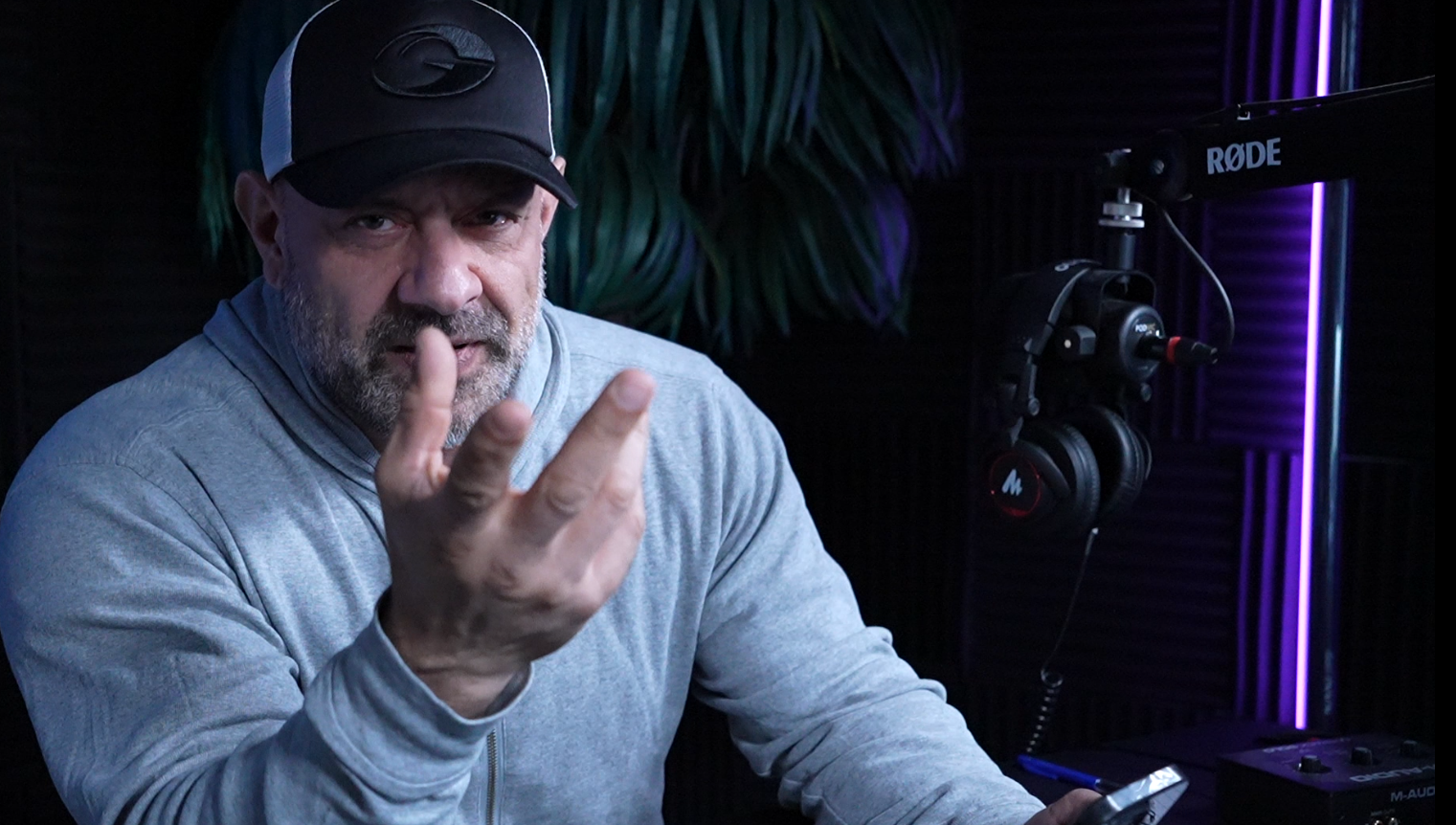 En man i en grå tröja och svart keps som sitter vid en mikrofonramp i ett ljudstudio eller podcaststudio, med hörlurar hängande. Mannen pekar med fingret mot kameran.