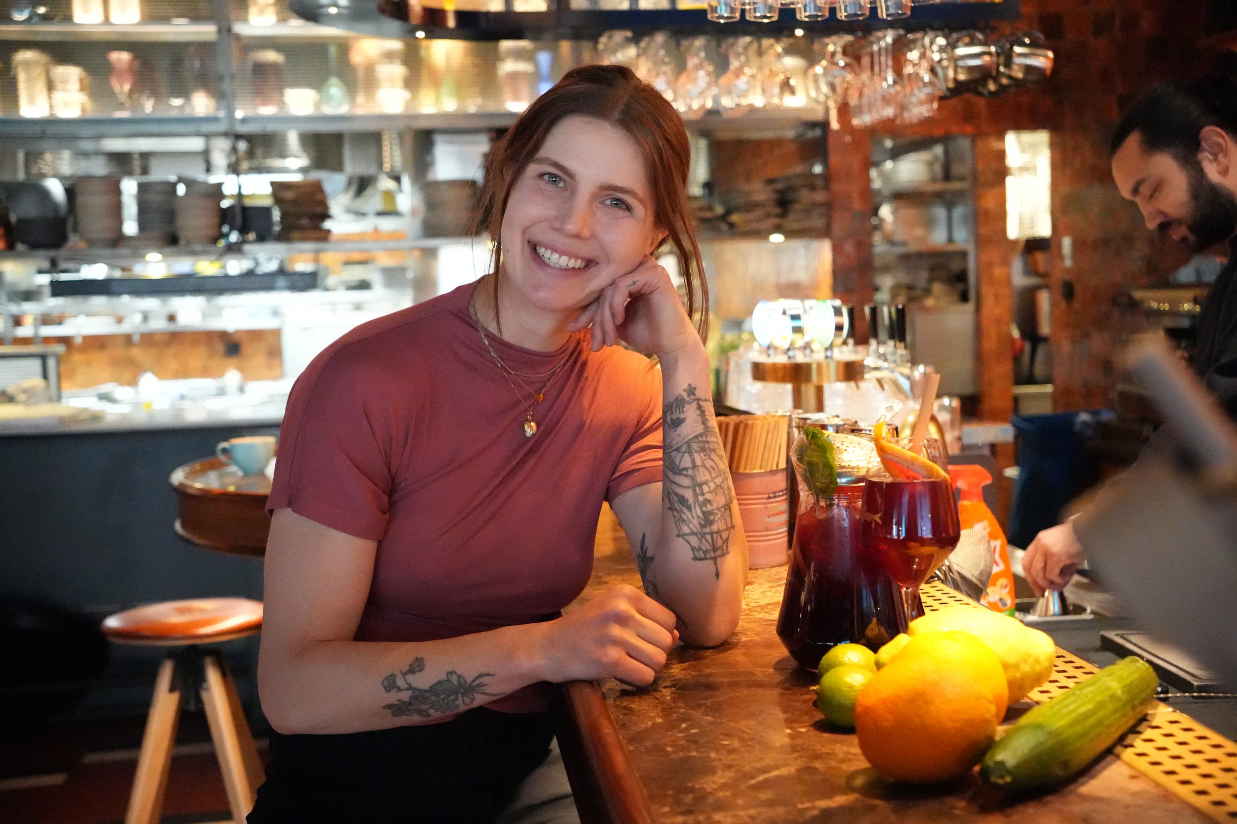 En kvinna med brunt hår och tatueringar på armen sitter vid en bar i en restaurang eller bar. Hon ler mot kameran, och framför henne finns ett glas cocktail, limetter och citrusfrukter. Bakgrunden visar stängda köksskåp och glasögon.