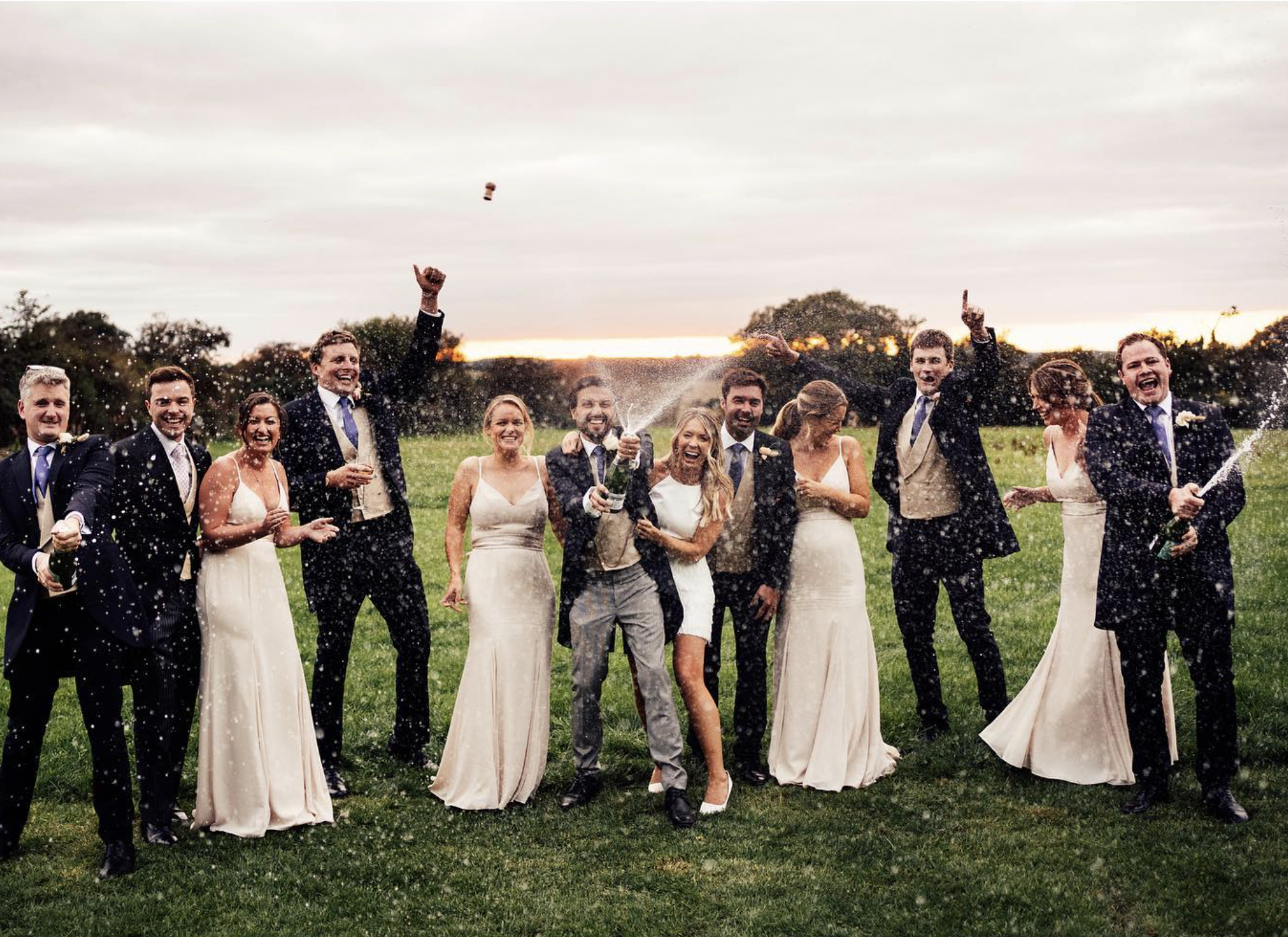 A group of wedding guests celebrating outdoors, popping a bottle of champagne, with a sunset in the background.