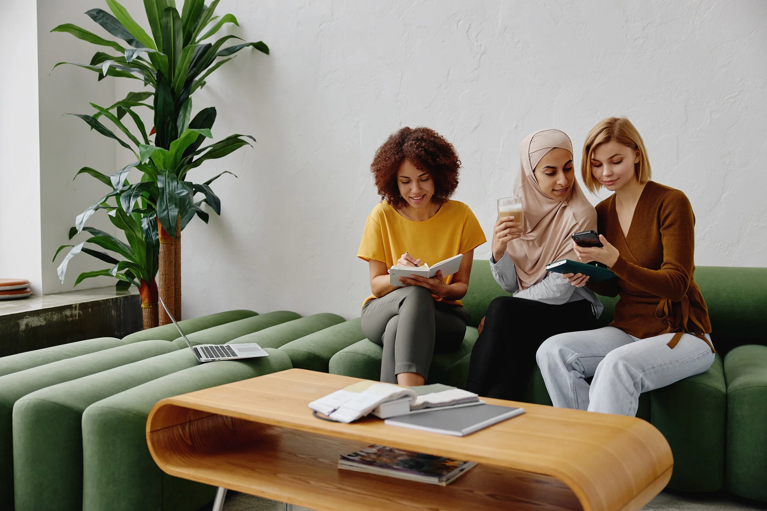 Three women sitting on a green sofa in a modern living room, engaged with their phones and notebooks, with a large potted plant nearby, a laptop on the table in front of them, and a white textured wall in the background.