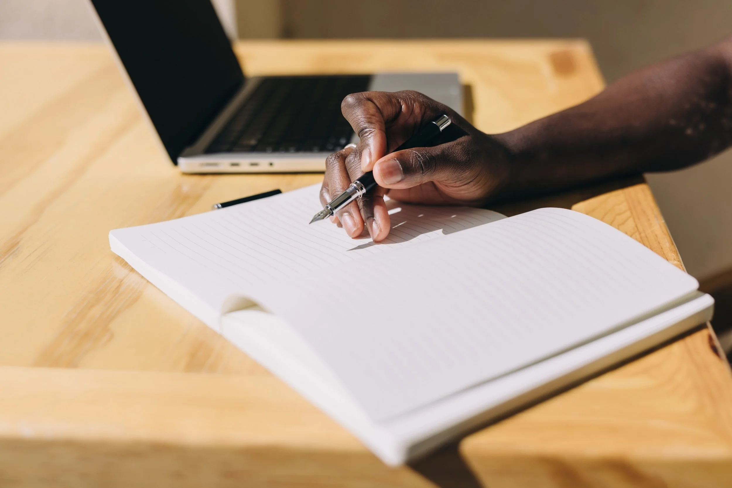 A hand holding a black pen over an open lined notebook on a wooden desk, with a laptop in the background.