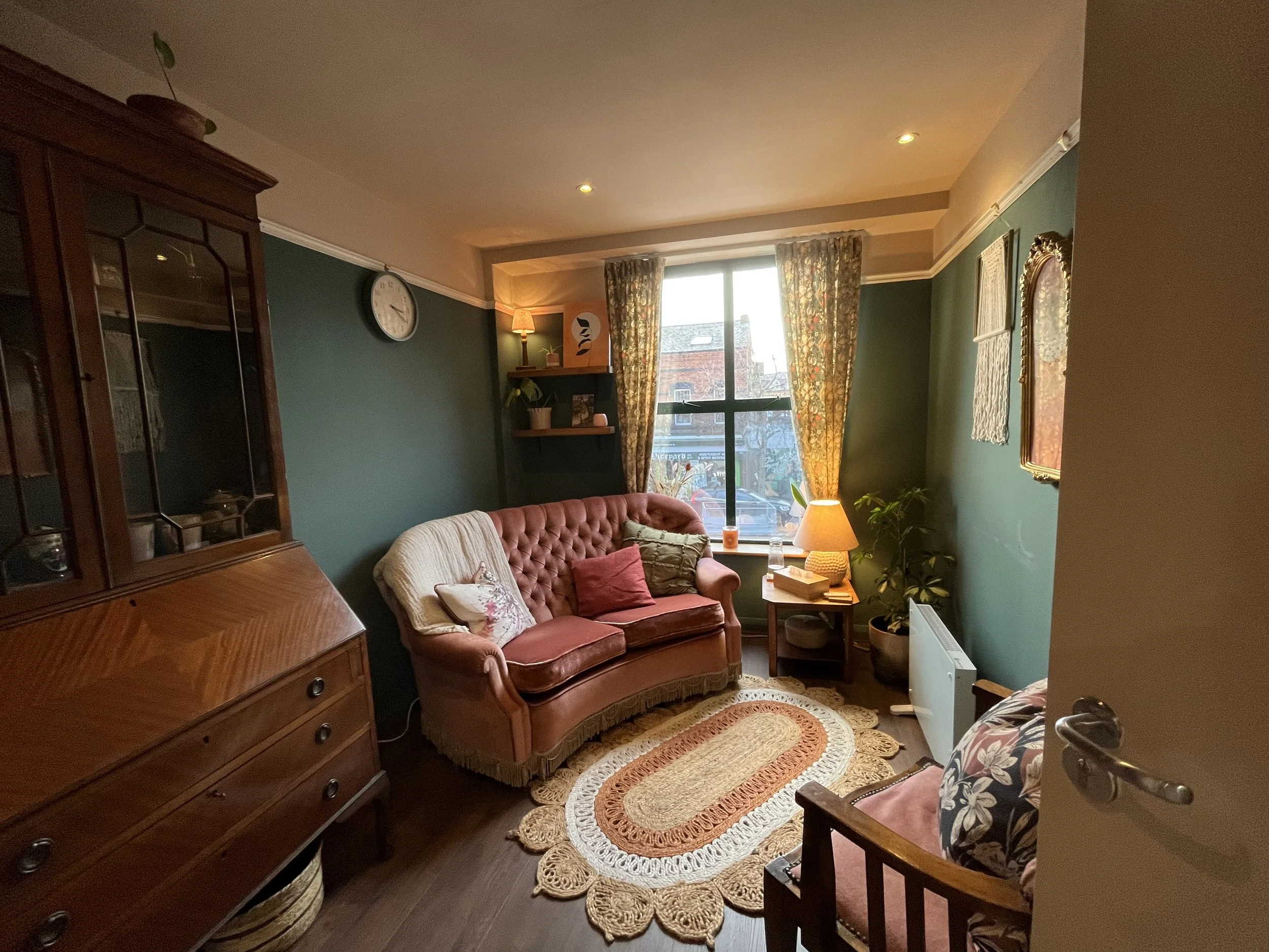 A cozy living room with a green wall, a floral curtain over a large window, a vintage pink velvet tufted sofa with pillows, a small side table with a lamp, a plaid armchair, a round woven rug, a wooden cabinet, and potted plants.