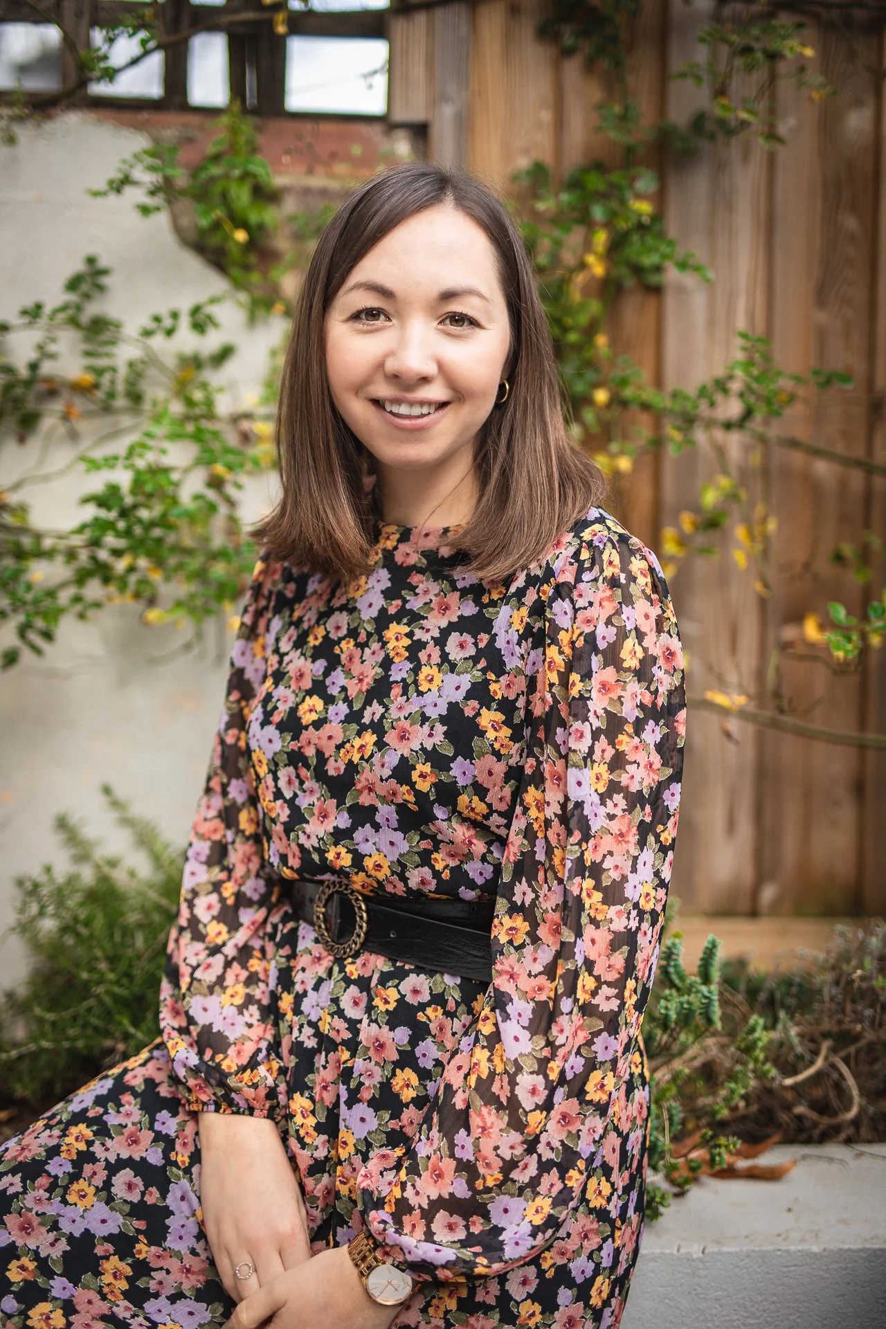A woman smiling outdoors, wearing a floral dress and a wristwatch, with greenery and a wooden fence in the background.