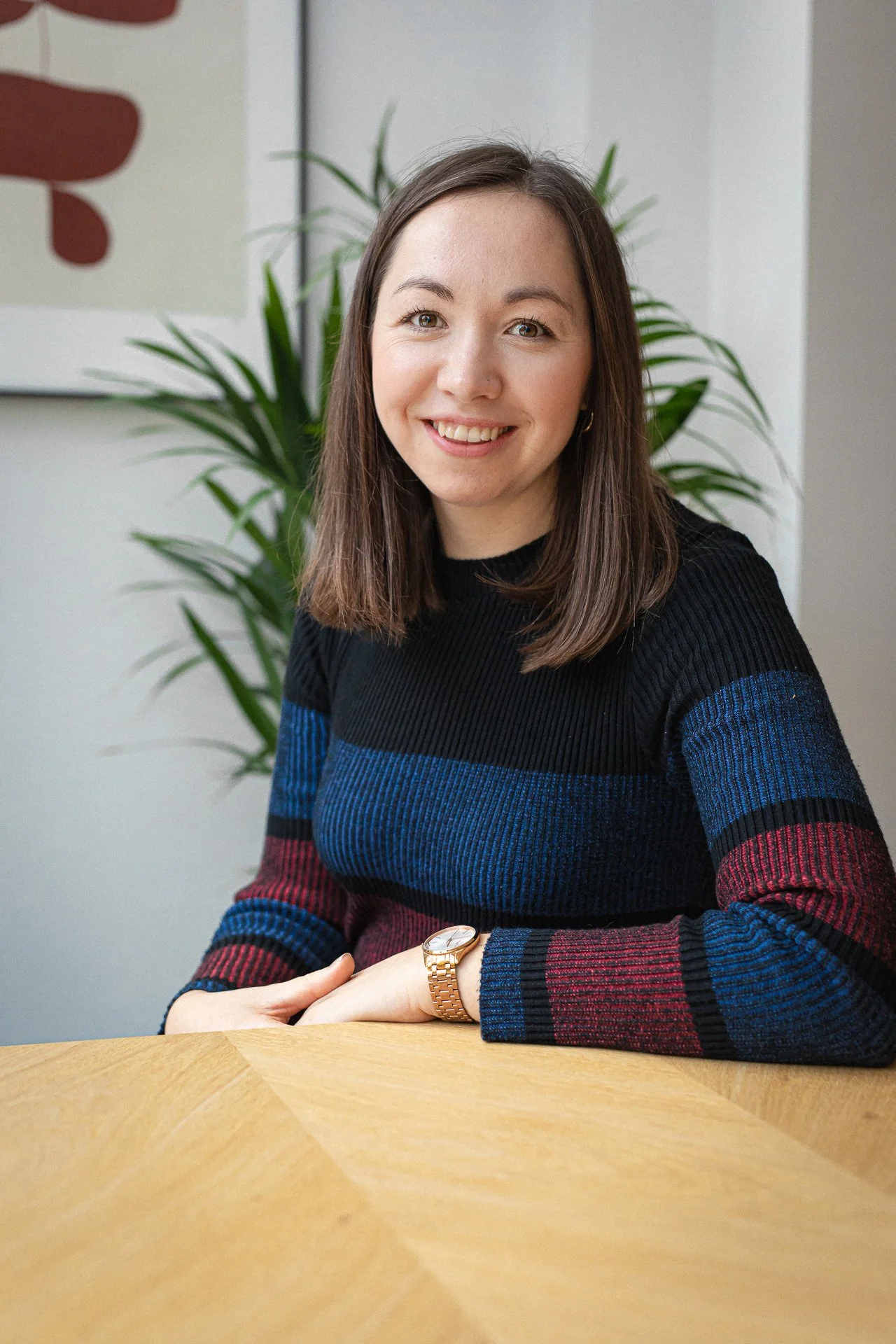A woman with shoulder-length brown hair, wearing a colorful striped sweater and a gold wristwatch, seated at a wooden table with a large green plant and modern art in the background.