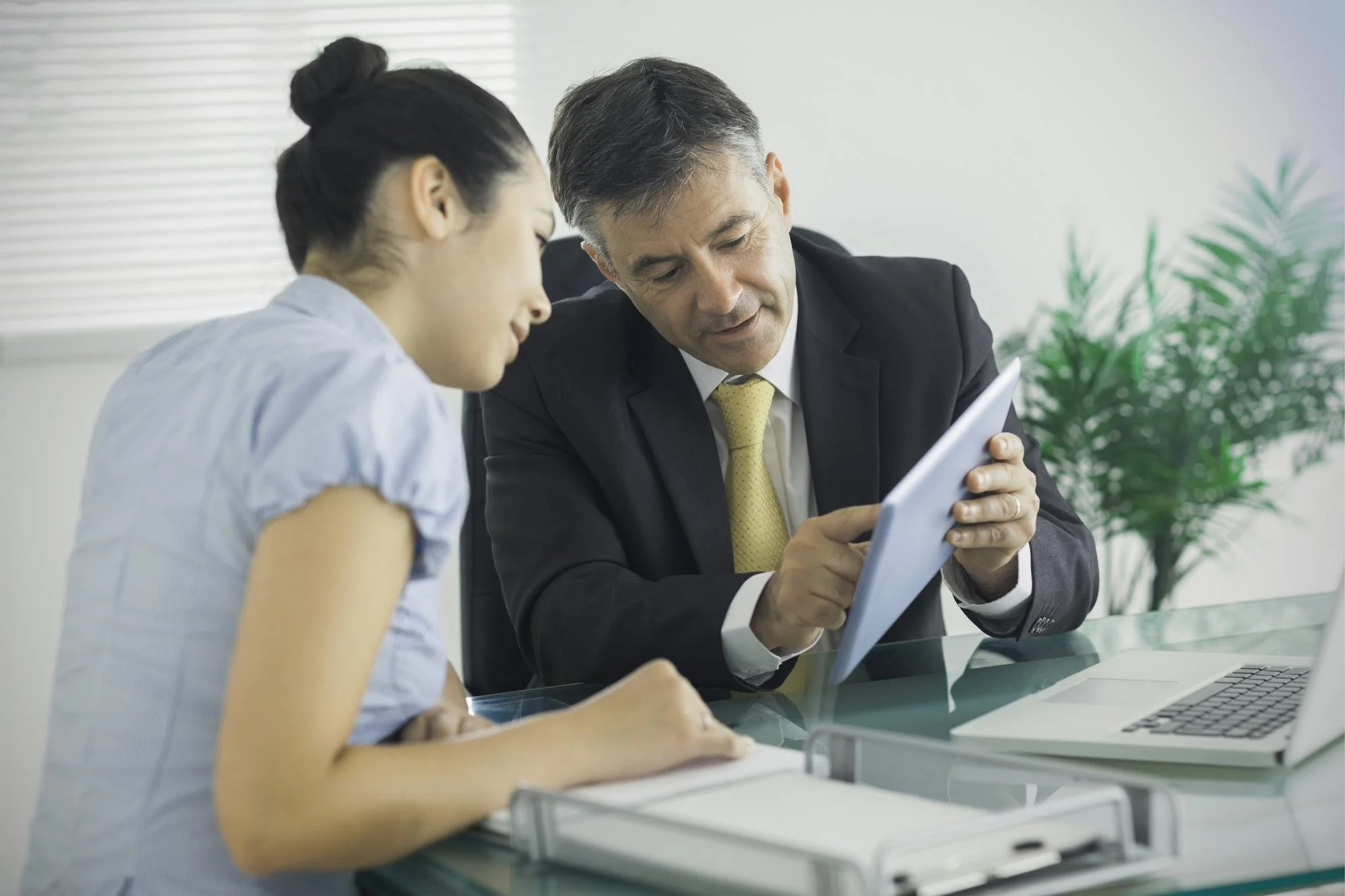 A man in a business suit showing a tablet device to a young girl in a school uniform at a glass table in an office setting.