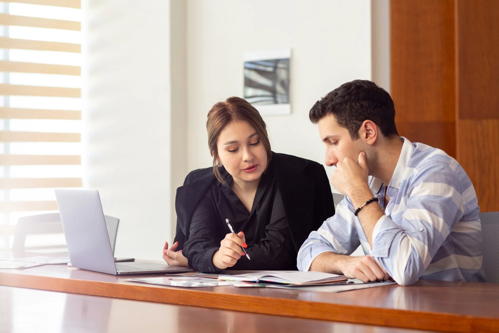Two young adults, a man and a woman, are sitting at a desk in a professional setting, engaged in a discussion with documents and a laptop in front of them.
