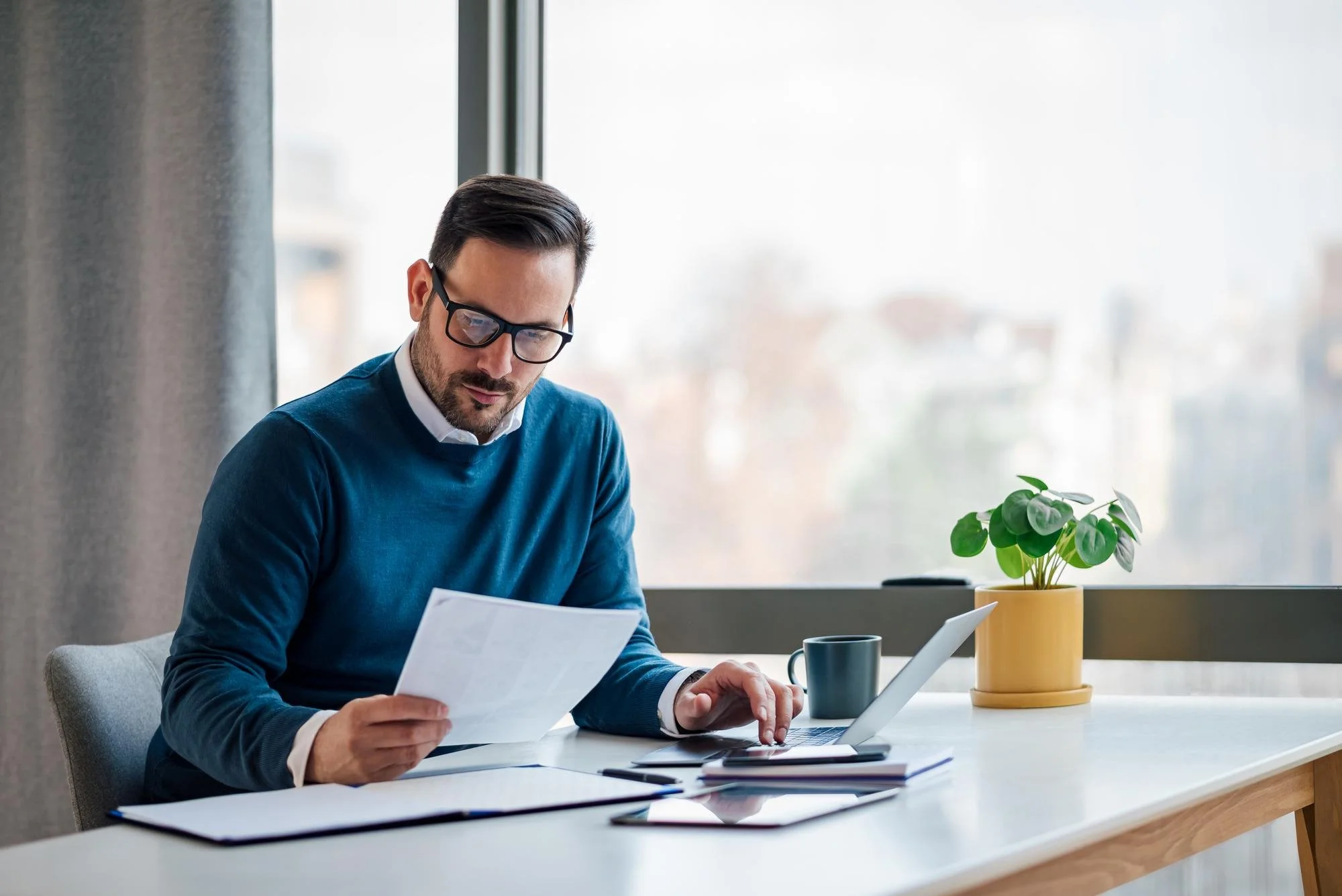A man with glasses and dark hair, dressed in a blue sweater, sitting at a white table, reading documents with a laptop, a coffee mug, and a tablet in front of him, in a bright office with large windows and a potted plant.