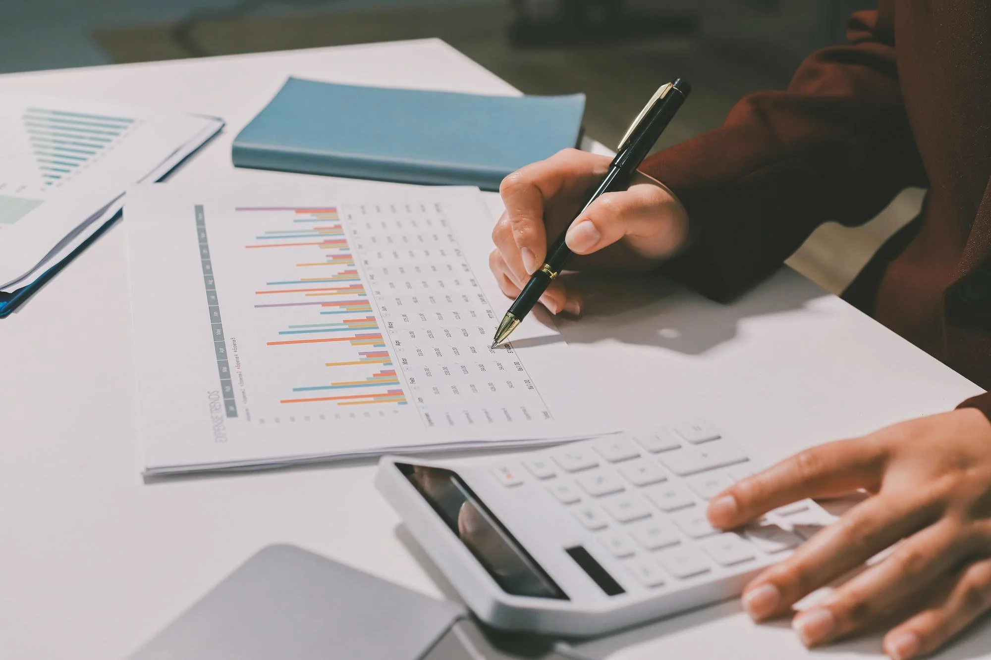 Person using a calculator and writing on financial documents with charts and graphs on a desk.