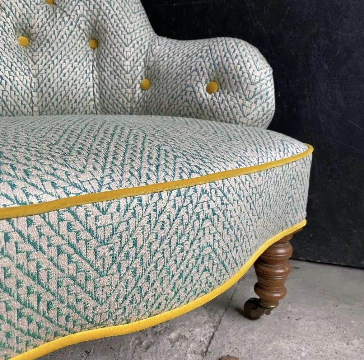 Close-up of an upholstered vintage chair with patterned fabric in green and pink, yellow piping, and turned wooden legs, set against a dark background.