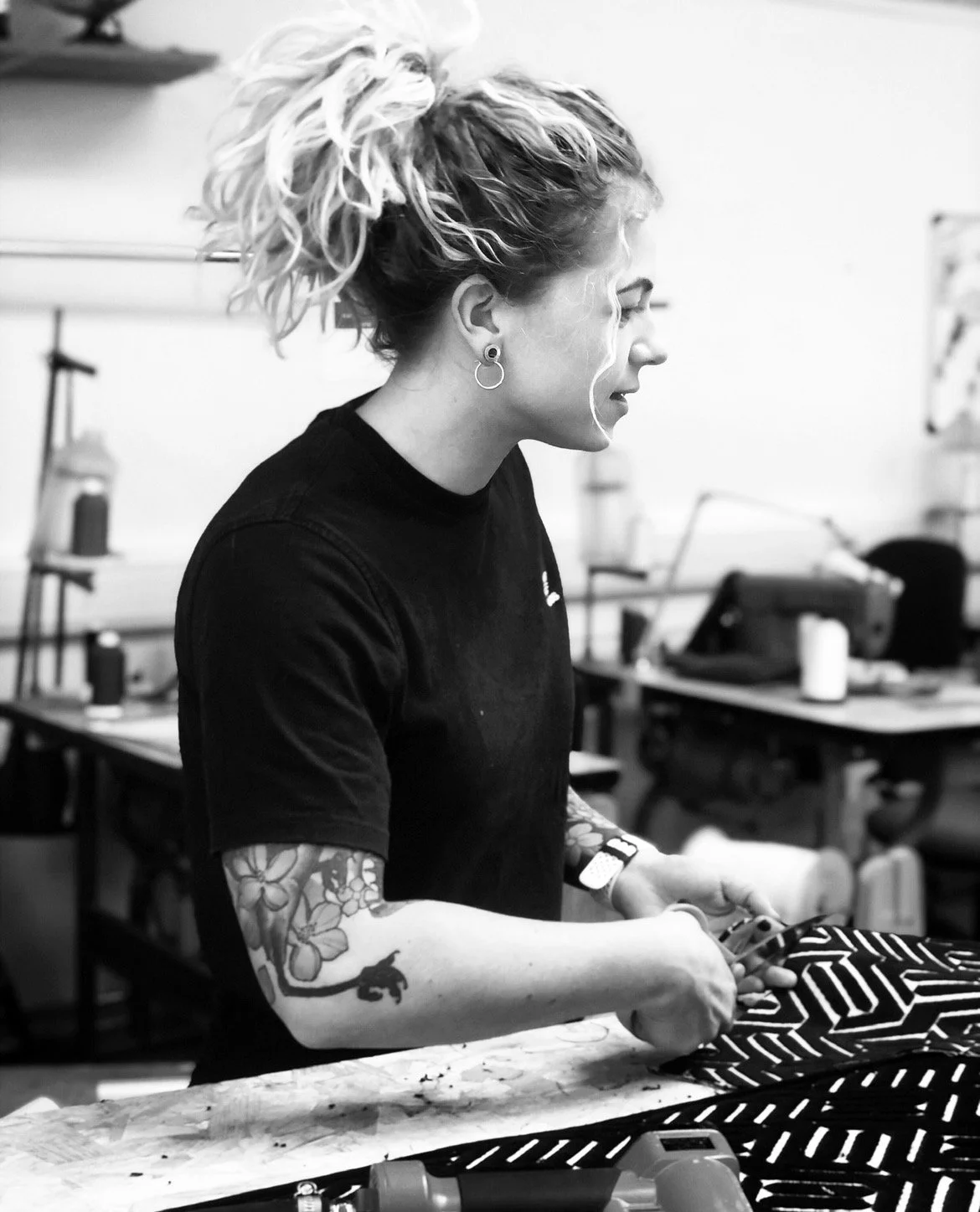 A woman with curly blonde hair in a ponytail, wearing earrings and a black shirt, is working with fabric in a workshop.
