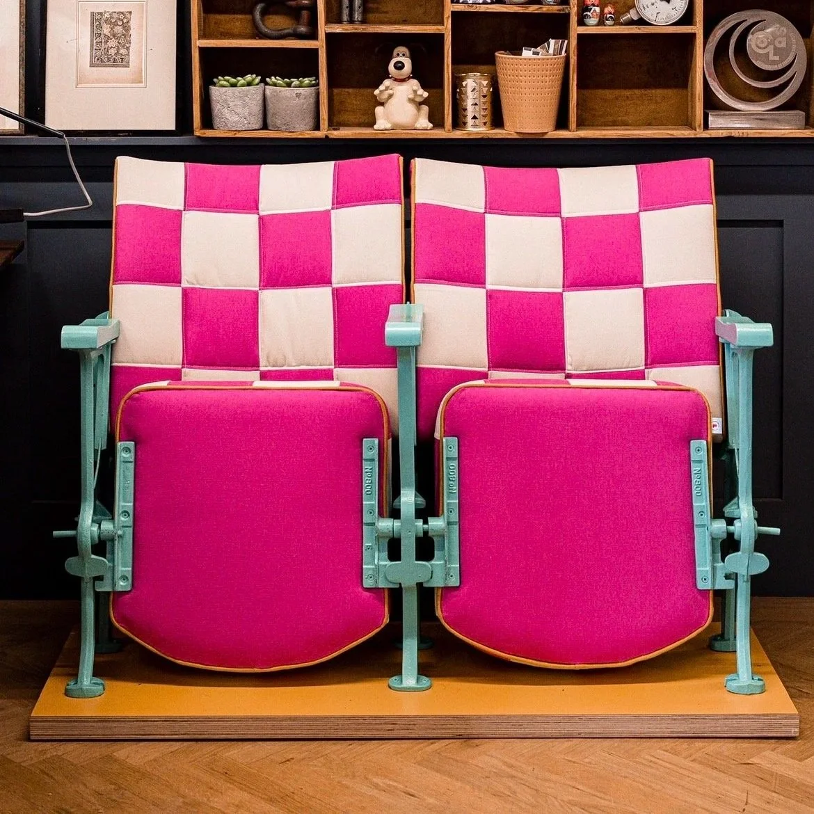 A pink and cream checkered vintage theater seat with mint green metal armrests, placed on a wooden platform in front of a dark wall with wooden shelving containing various decor items, including plants, sculptures, and books.