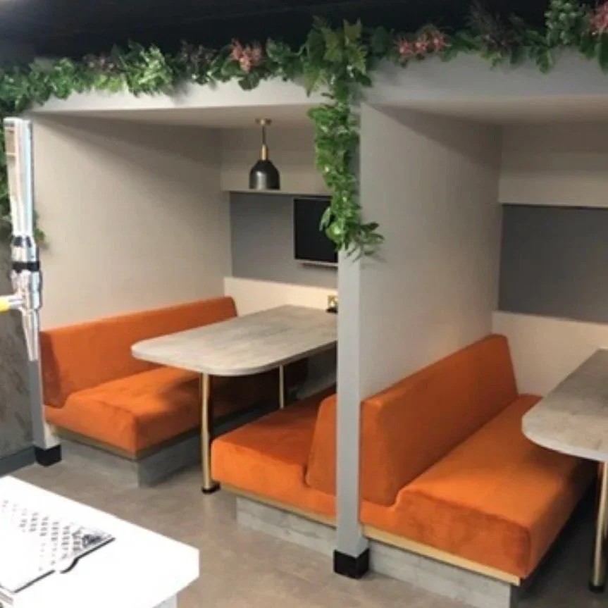 Two adjacent booth-style restaurant seating areas with orange cushioned benches, separated by a half wall with green plants on top, and a marble table. There is a wall-mounted TV and a black pendant light in the background.