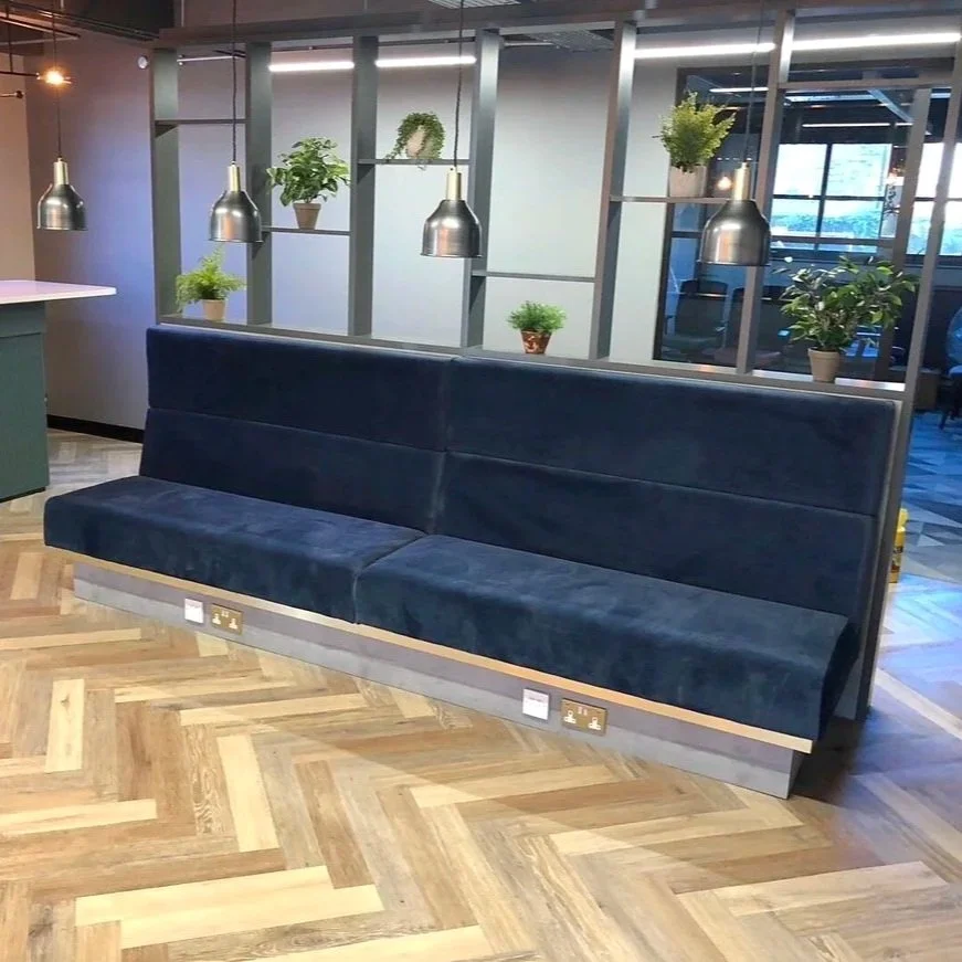 A modern office lounge area with a dark blue cushioned bench against a decorative black metal partition filled with potted plants. The floor is wooden with a herringbone pattern, and there are hanging metallic pendant lights and a nearby wall with wi