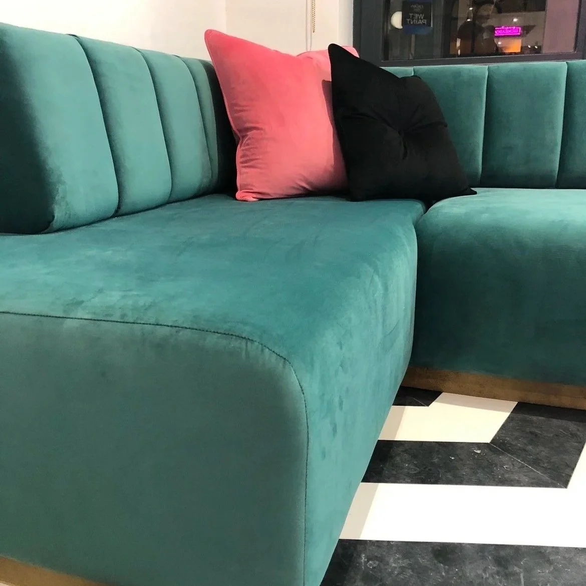 A teal sectional sofa with three cushions in pink, black, and teal in a modern interior. The floor has a black and white geometric pattern.