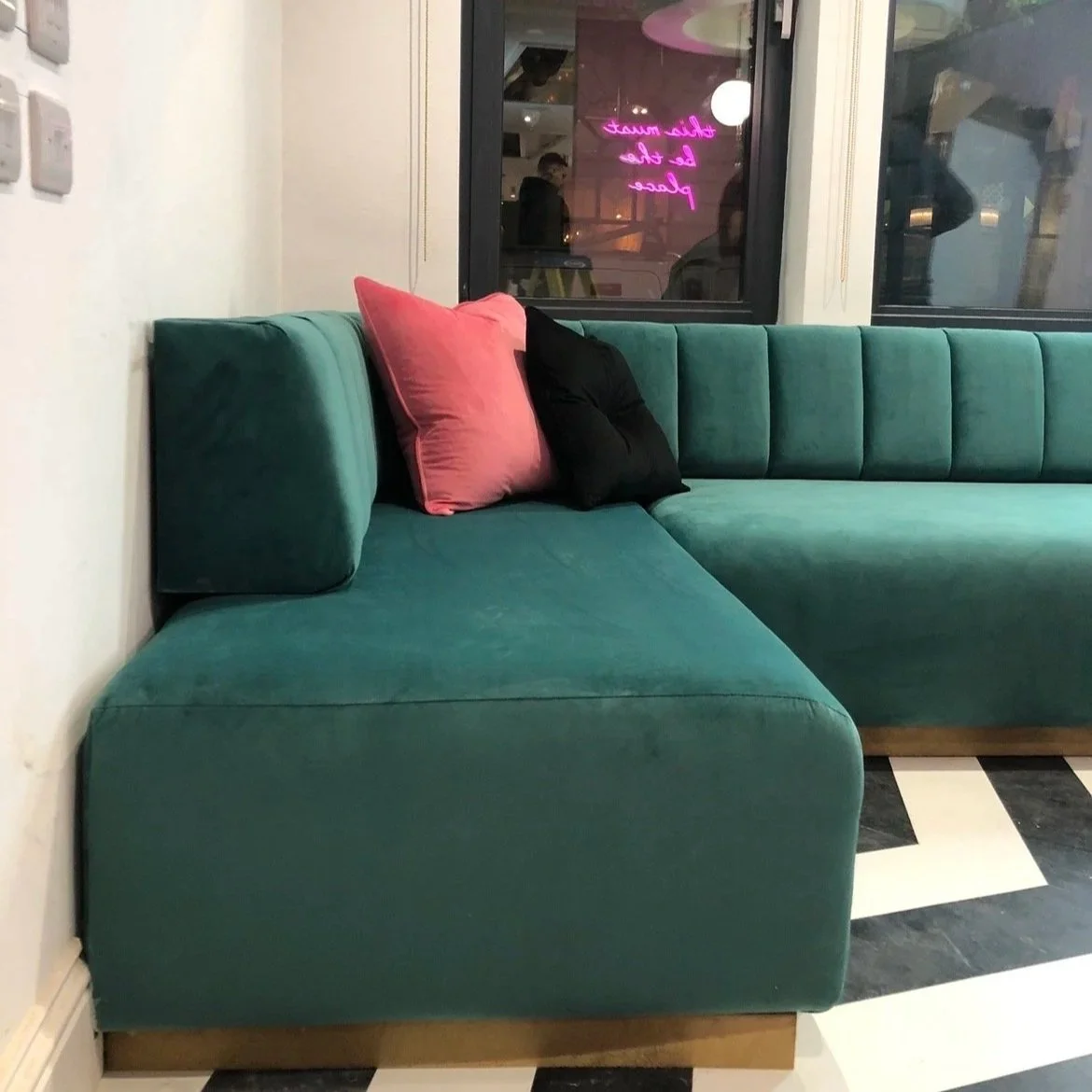 A green upholstered sectional sofa with pink and black throw pillows, sitting in a room with black and white checkered flooring and large windows. Neon sign visible through window, reflecting pink word.