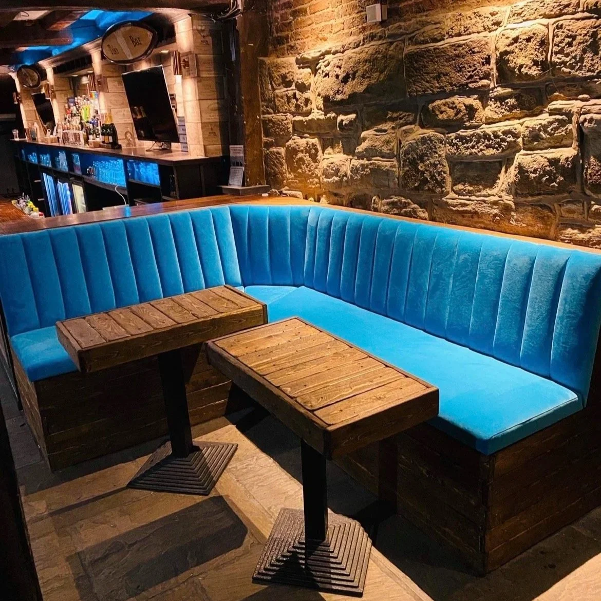 A cozy bar area with a curved blue velvet booth, two small wooden tables, and a stone wall background.