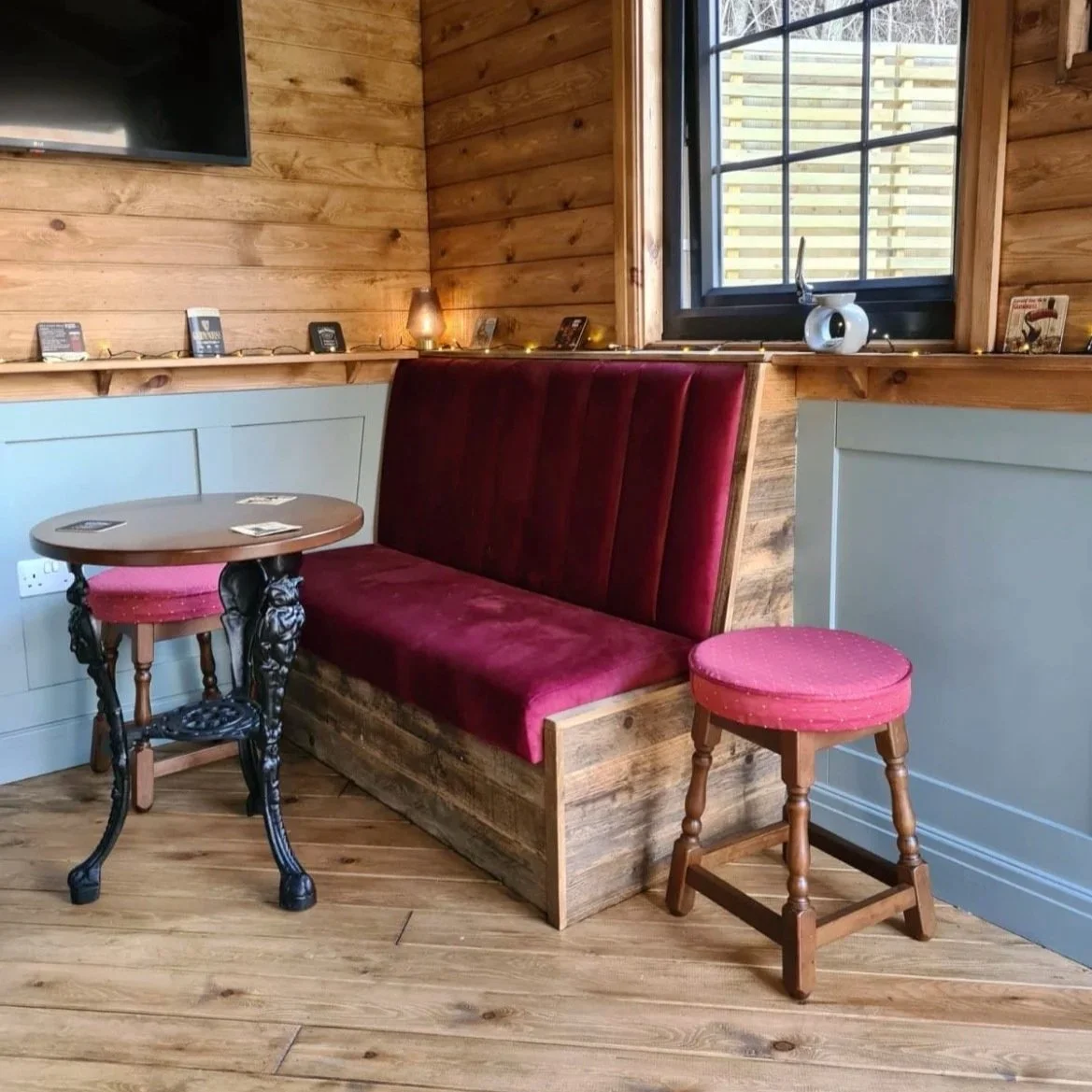 A cozy corner with wooden walls and floor, featuring a red upholstered bench seat with a wooden frame, a small round table with ornate black legs, a pink cushioned stool, a small white ceramic teapot on the windowsill, and a string of yellow fairy li