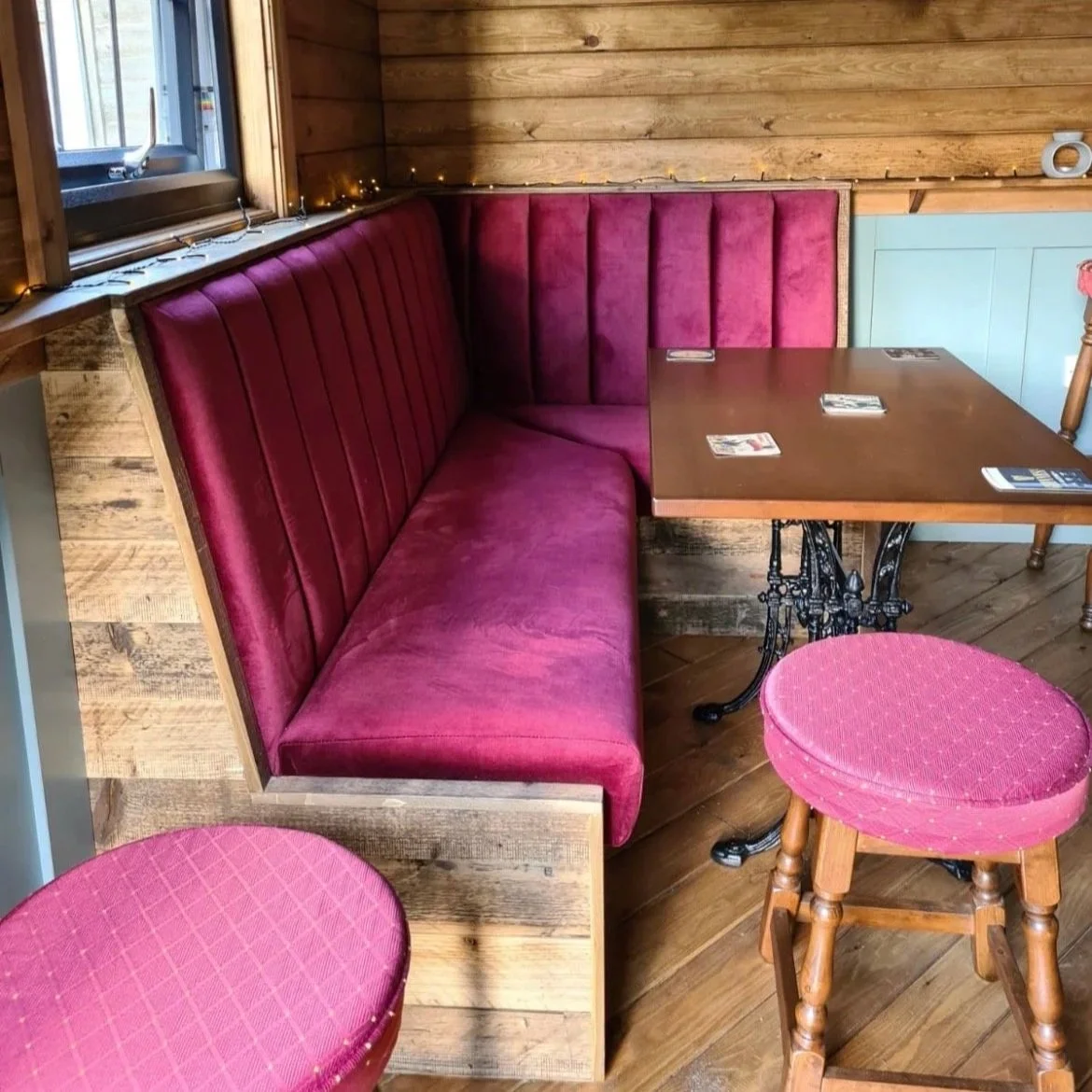 A seating area with a red velvet booth, a wooden table, and pink upholstered stools in a rustic restaurant setting.