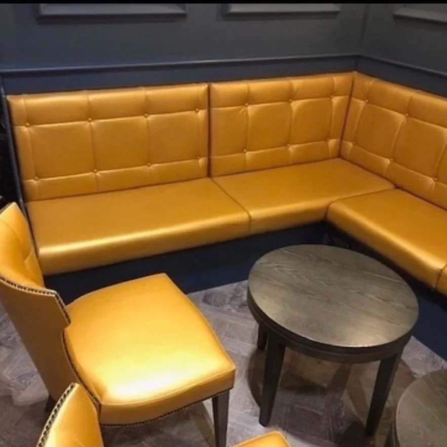 Corner of a room with mustard yellow leather seating, including a sectional sofa and chairs, and dark wood round tables on a gray patterned carpet.