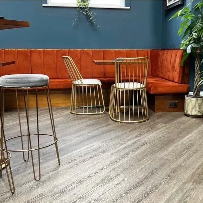 A corner of a modern restaurant or cafe with a blue wall, orange upholstered bench seating, a small round table, metal wire chairs, a tall metal stool with a cushioned seat, and a large plant in a decorative pot.