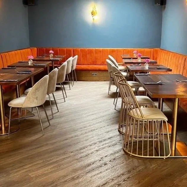 An empty restaurant with wooden tables, beige chairs, and a long orange banquette along a teal wall. Small vases with pink flowers are on each table.