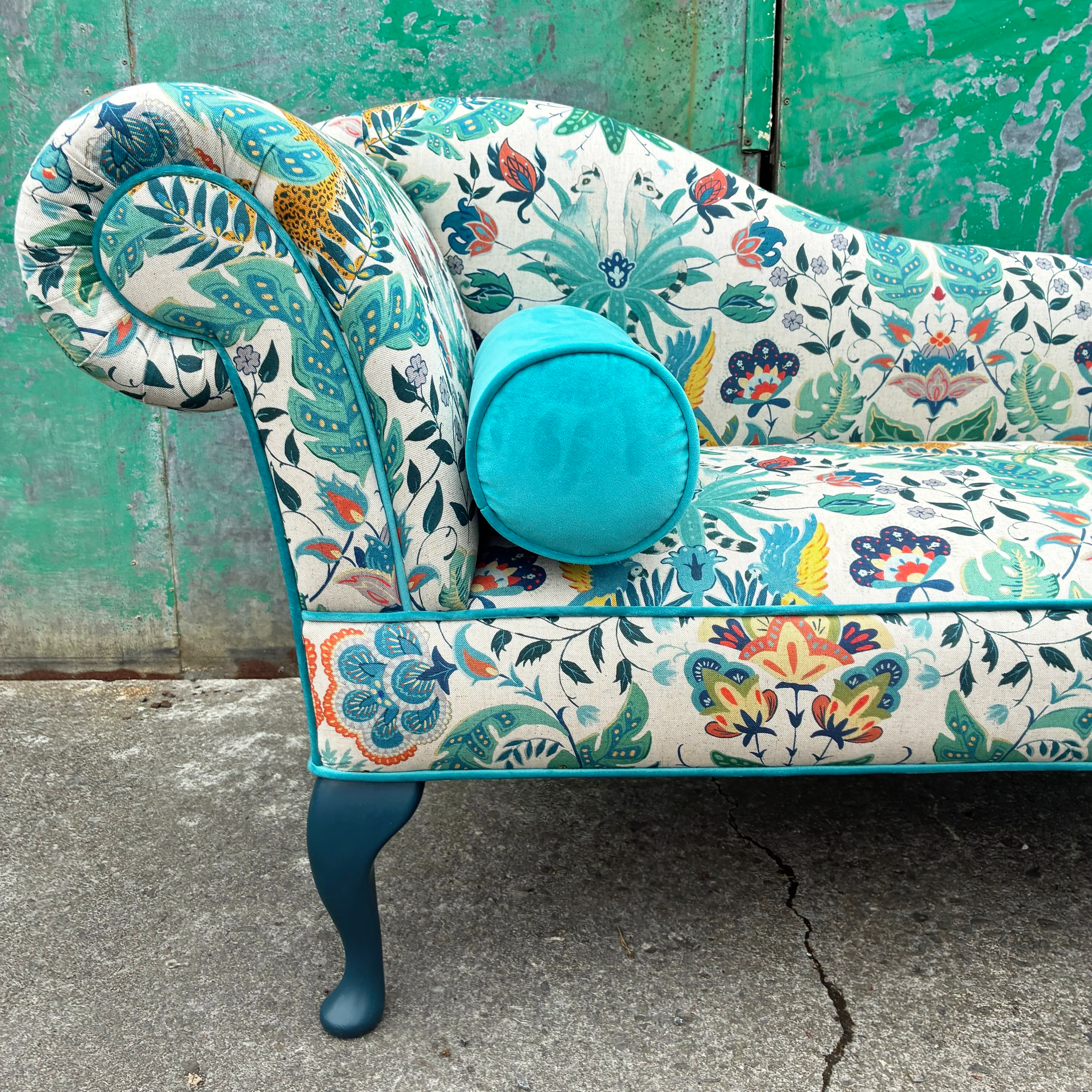 A colorful floral-patterned sofa with matching pillows and a bright turquoise oval ottoman, placed on an outdoor concrete surface against a weathered green door.
