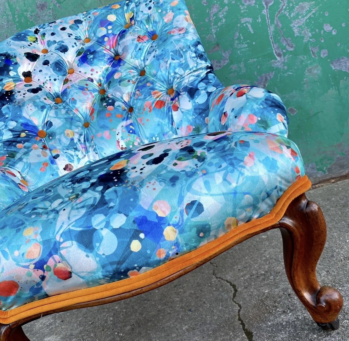 A vintage armchair with a colorful, abstract, blue, and orange patterned fabric upholstery and wooden legs, situated beside a green, distressed wall on a concrete surface.