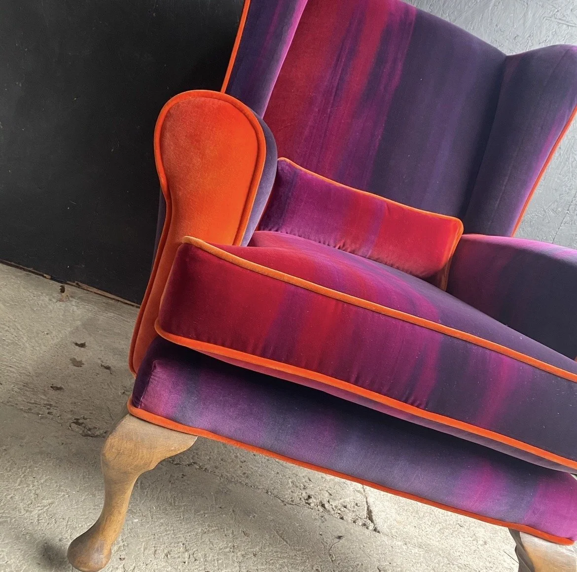 Close-up of a vintage purple velvet sofa with orange piping and a matching orange velvet armrest, wooden carved legs, set against a concrete floor and wall.