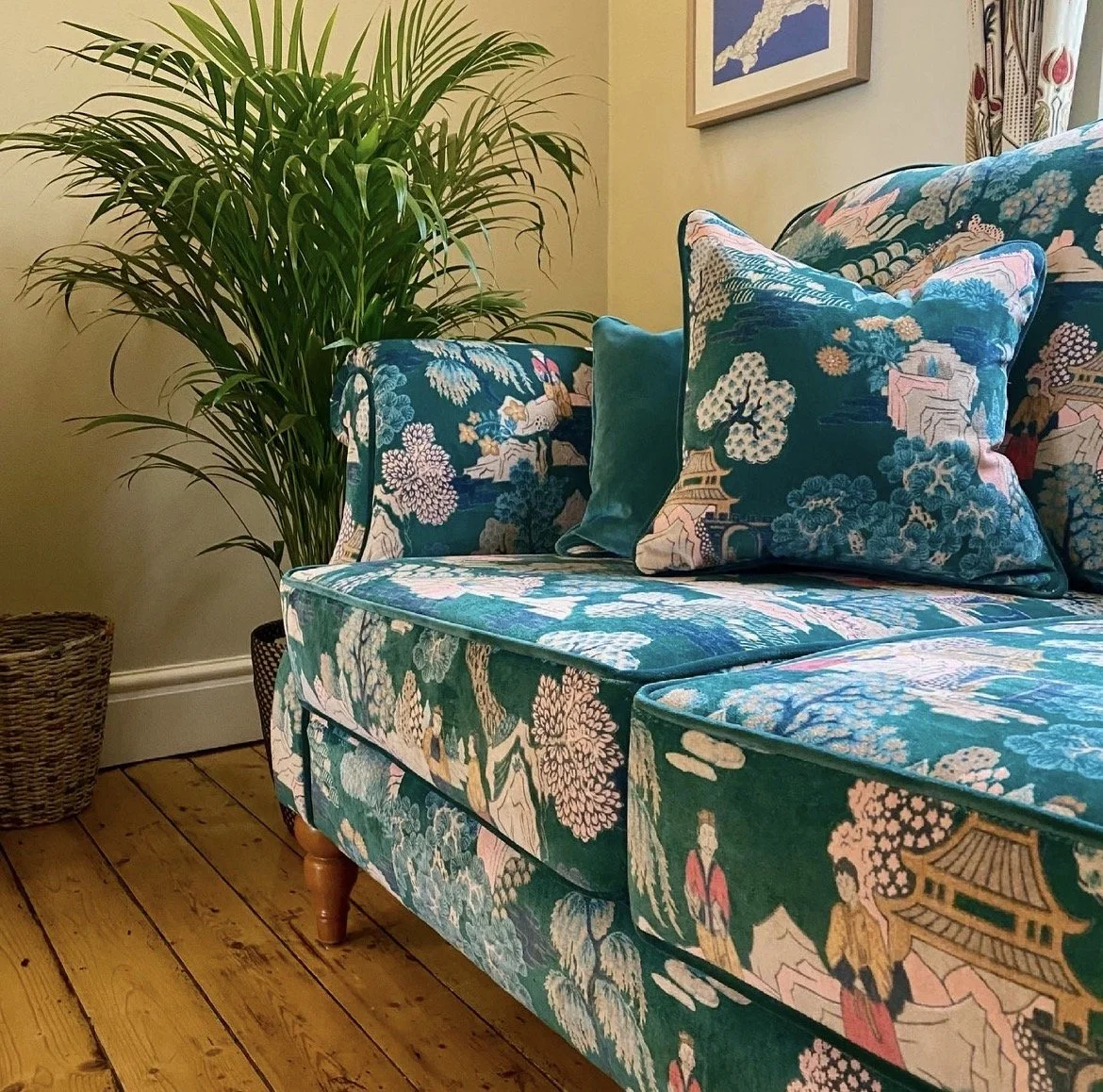 A living room corner with a floral-patterned sofa and cushions, a large indoor plant, a wicker basket, hardwood flooring, and wall art.