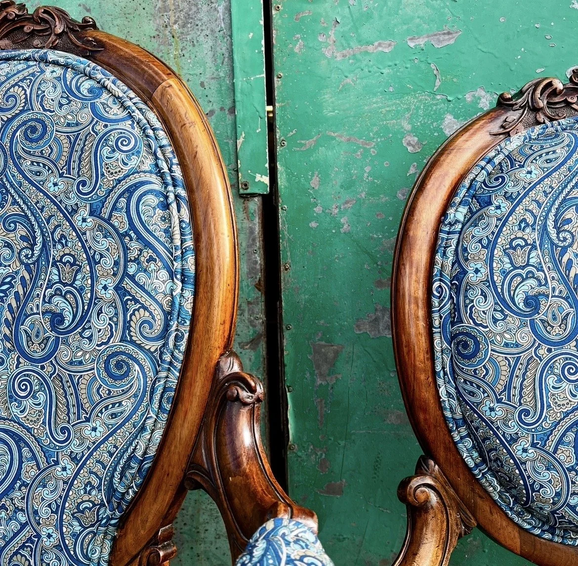 Two vintage wooden armchairs with ornate carvings and blue patterned fabric upholstery, placed in front of a green, weathered wooden wall or door.