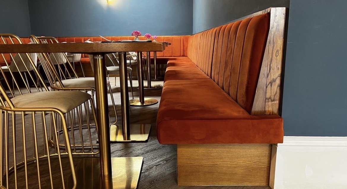 Restaurant interior with a long burnt orange velvet banquette, wooden wall paneling, and a table with gold and white chairs.