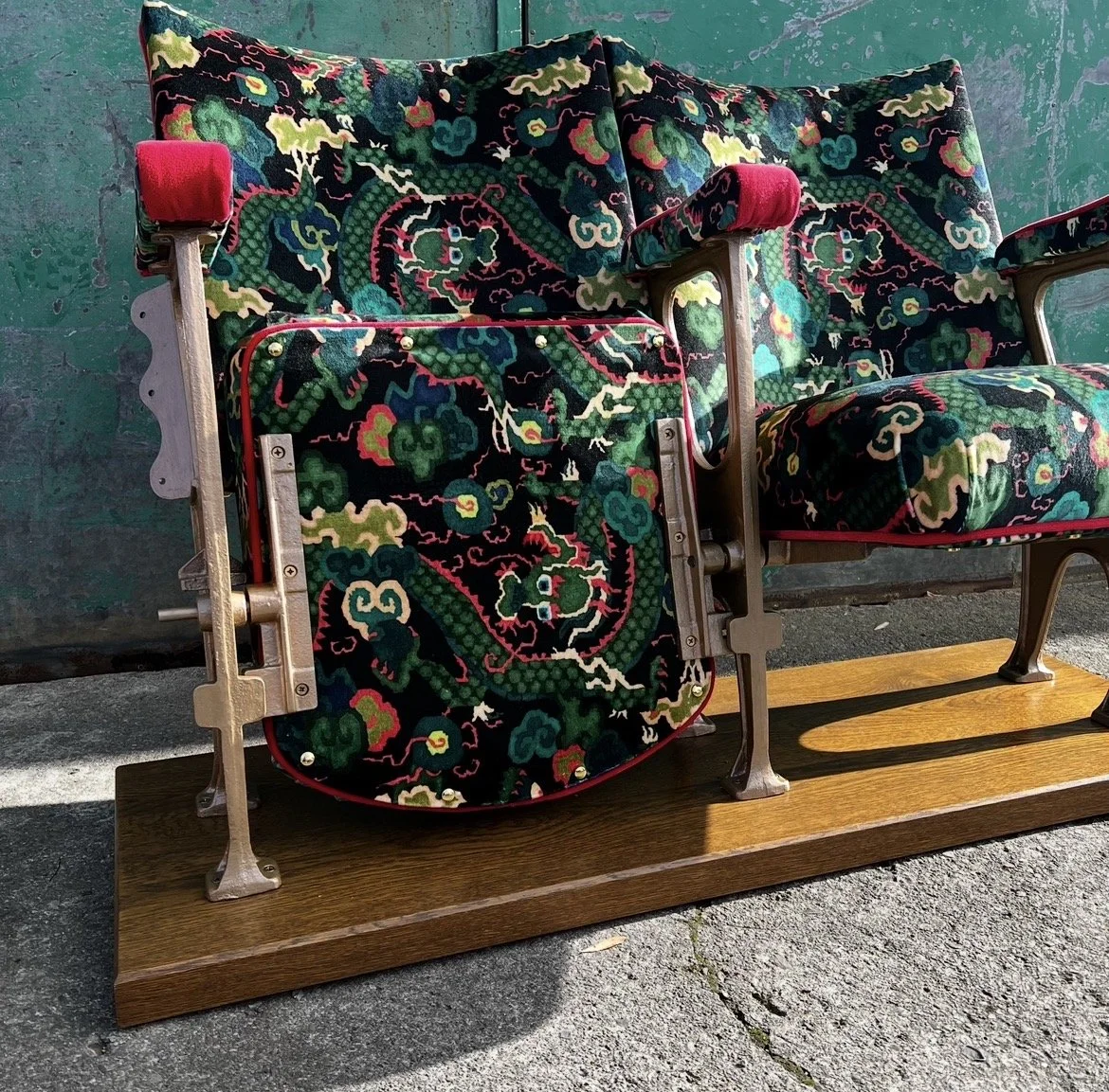 A vintage theater seat covered with colorful embroidered dragon design, with a wooden base and metal frame, bathed in sunlight.