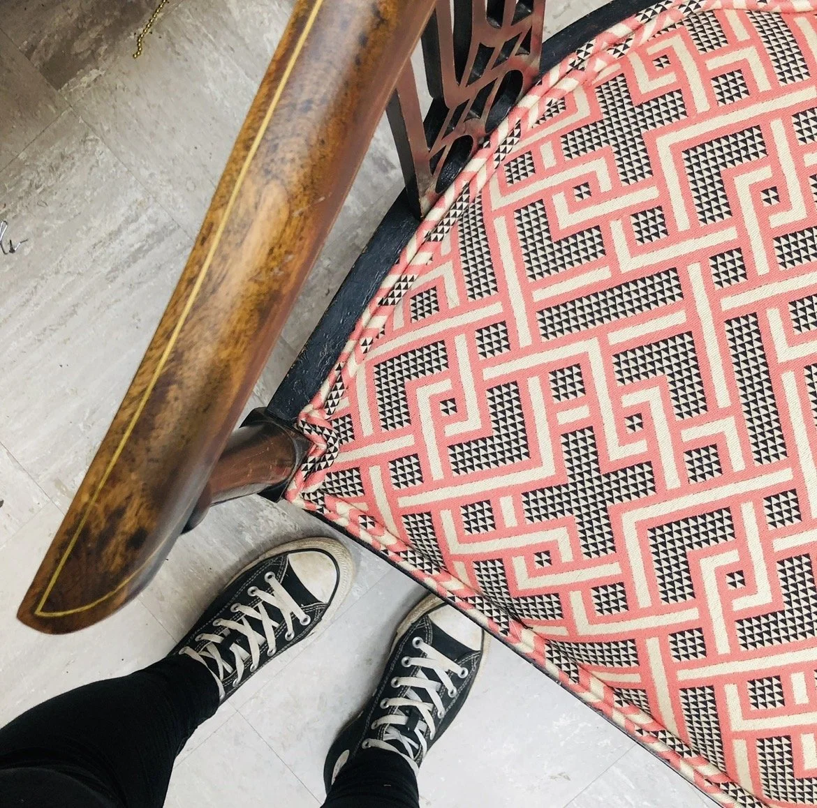 Top-down view of a person in black Converse sneakers standing on a light gray tiled floor, next to a wooden table with a patterned fabric overlay featuring pink, black, and white geometric shapes.