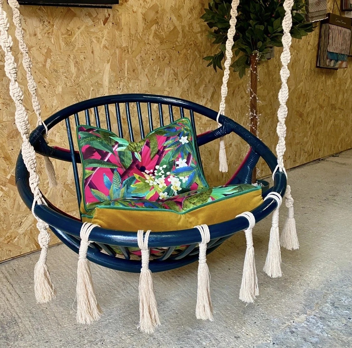 A round hanging chair with white tassels along the outer frame, positioned indoors against a wooden wall with a potted plant nearby. The seat has a colorful floral cushion.