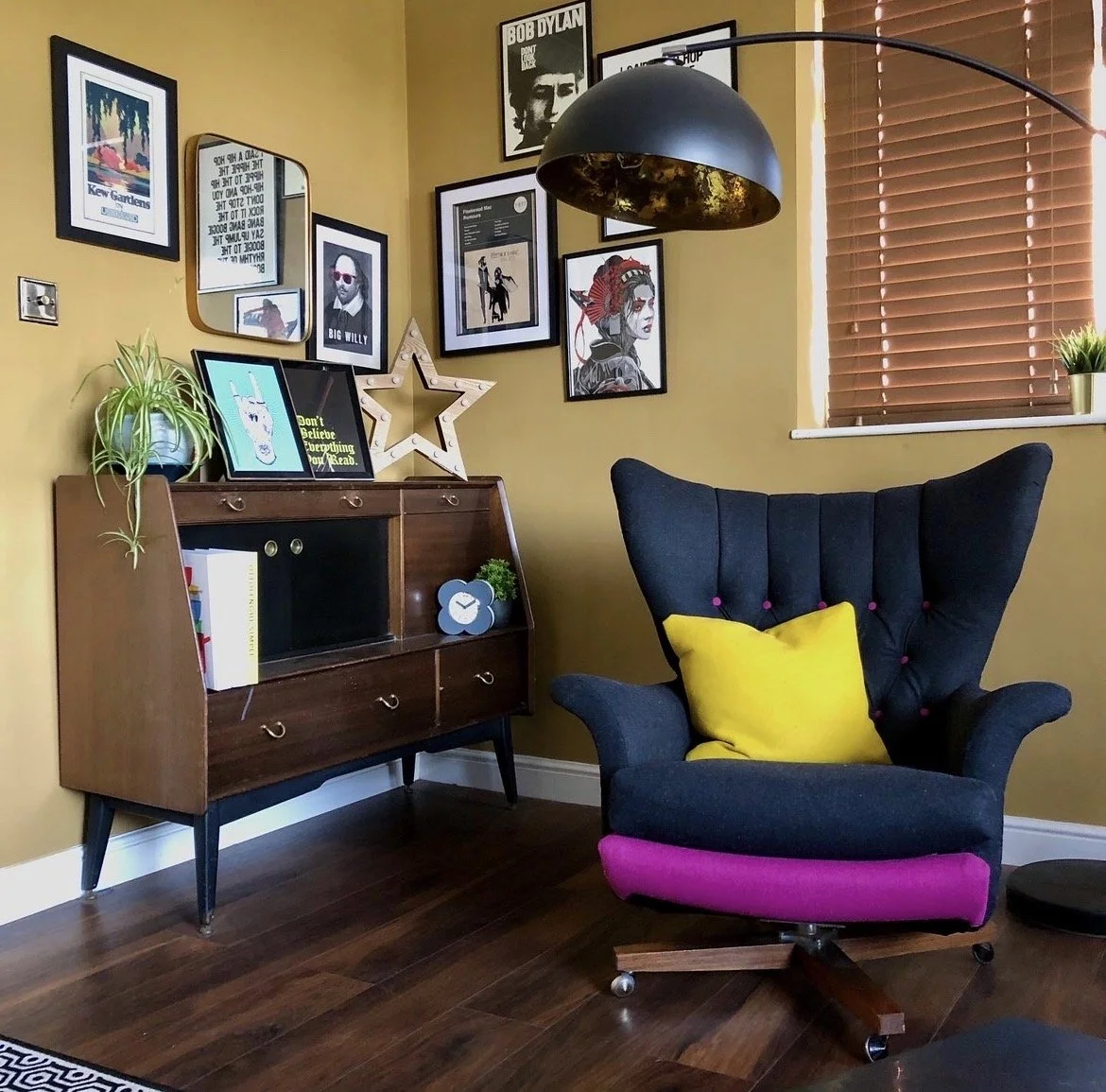 A cozy corner of a room with a dark armchair upholstered in black fabric with pink and yellow cushions. To the left is a vintage wooden cabinet decorated with framed artwork, a potted plant, and a star-shaped light. The wall behind features a gallery
