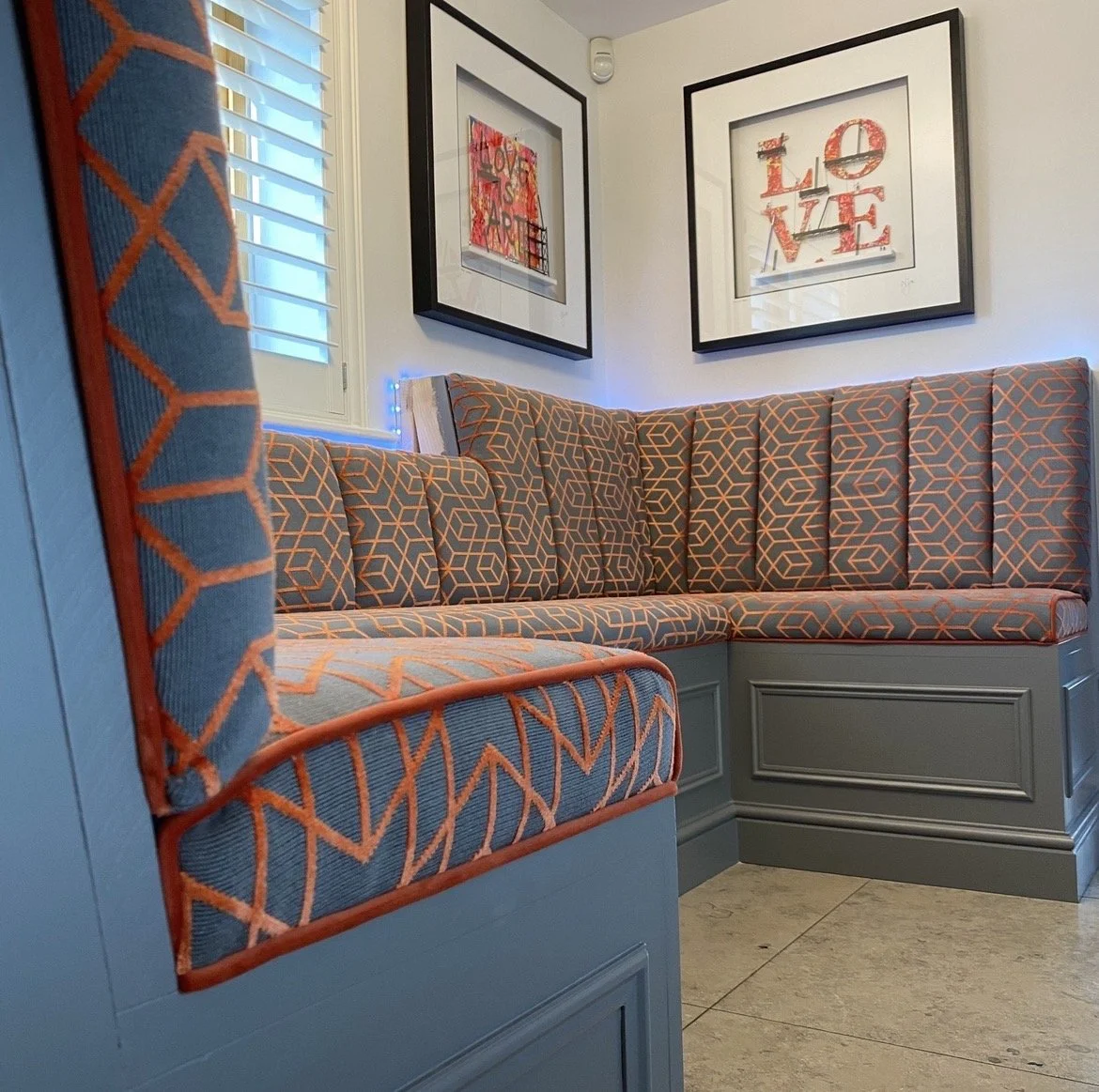 A corner of a room with a built-in blue and orange patterned cushioned bench, two framed art pieces on the white walls, one with the word 'LOVE' in red, and a window with shutters and string lights.