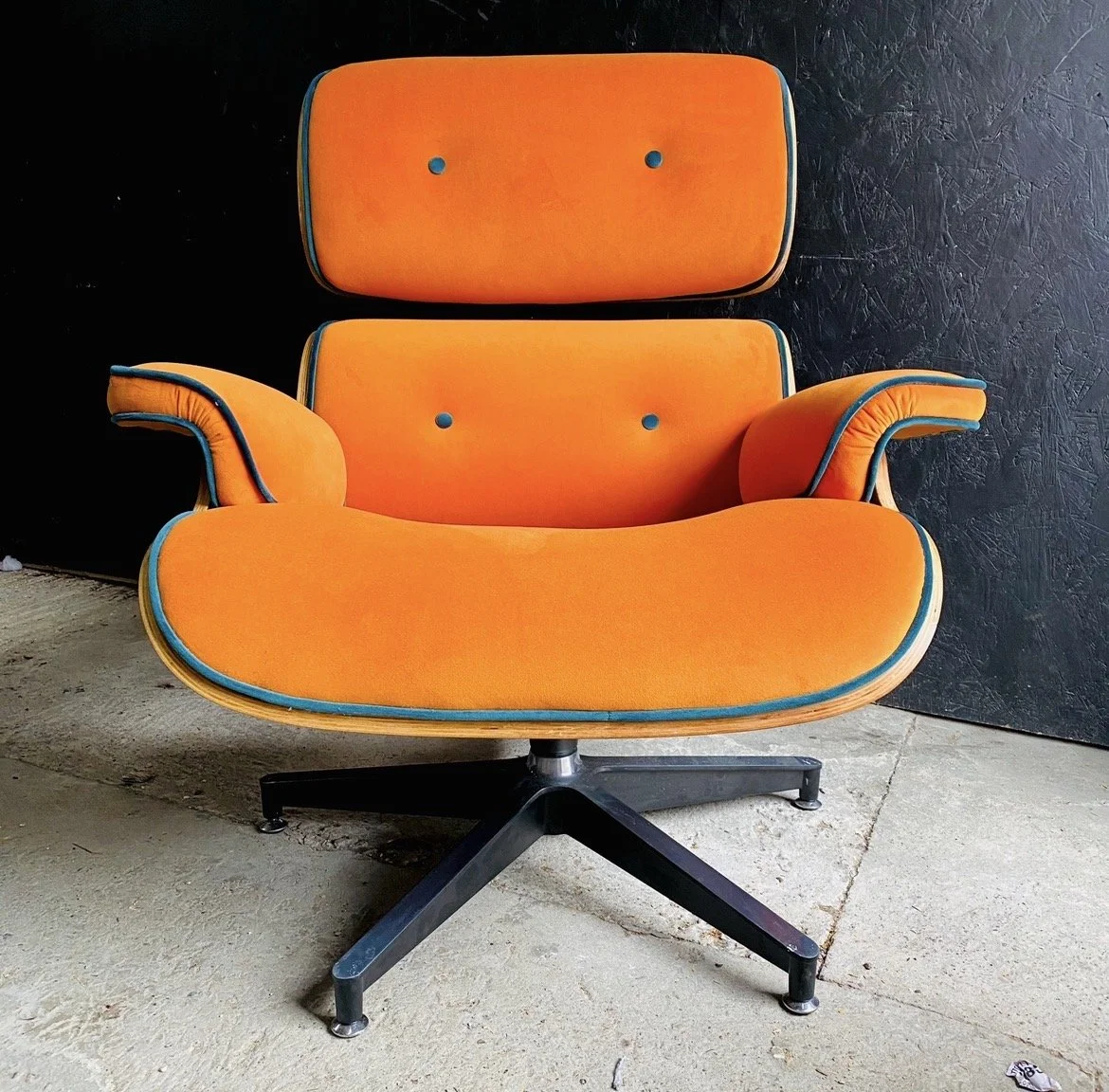 Orange upholstered armchair with a matching headrest, black metal base, and decorative blue piping, placed on a concrete floor against a black textured wall.