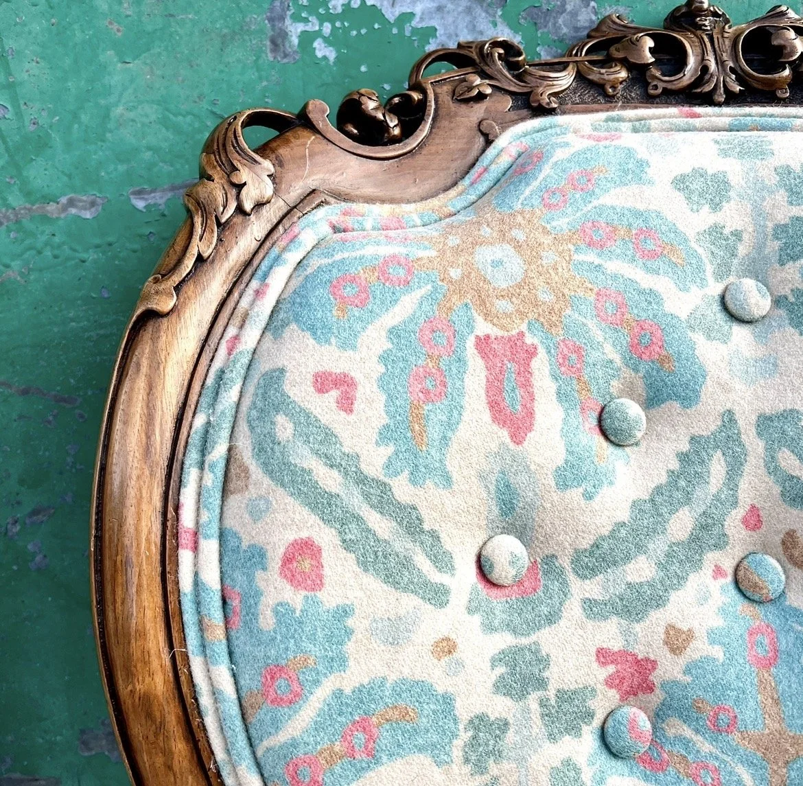 A close-up of a vintage upholstered piece of furniture, possibly a headboard or a chair, with a wooden carved frame and pastel patterned fabric. There are decorative fabric-covered buttons on the upholstery.