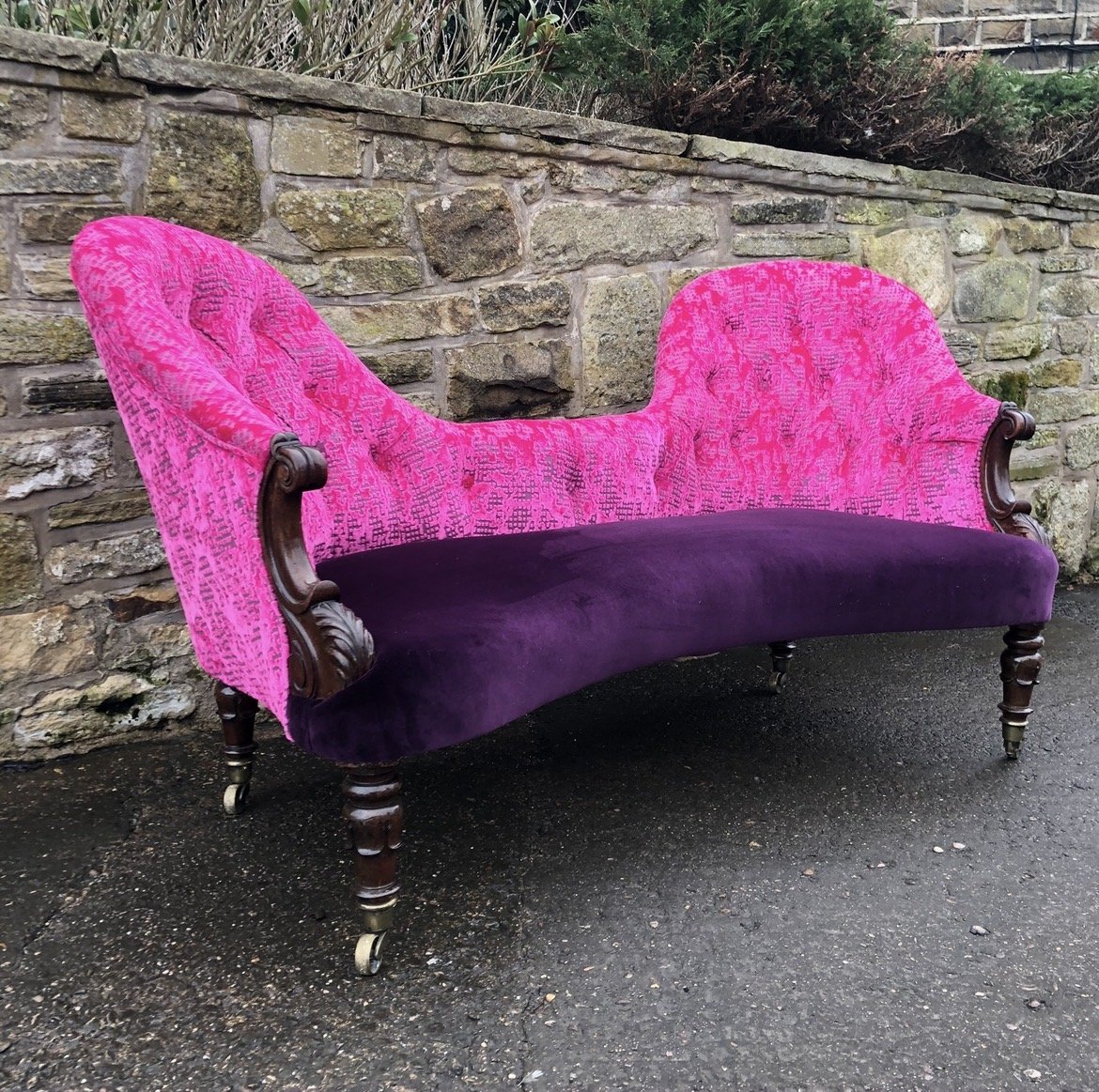 An antique Victorian style sofa with a pink patterned backrest and armrests, and purple velvet seat cushion, positioned outdoors against a stone wall.