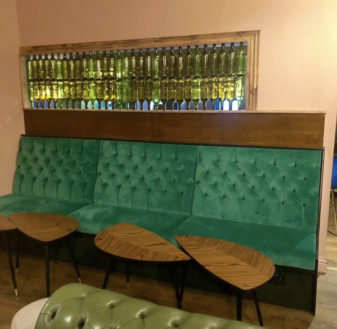 A green tufted booth seating with three wooden tables in front of it and bottles on shelves behind it.