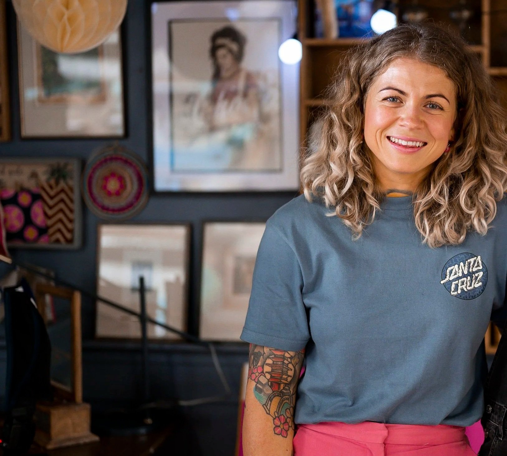A smiling woman with curly blonde hair, tattoos on her arm, wearing a blue Santa Cruz T-shirt and pink pants, standing in an art studio with framed pictures and artwork behind her.