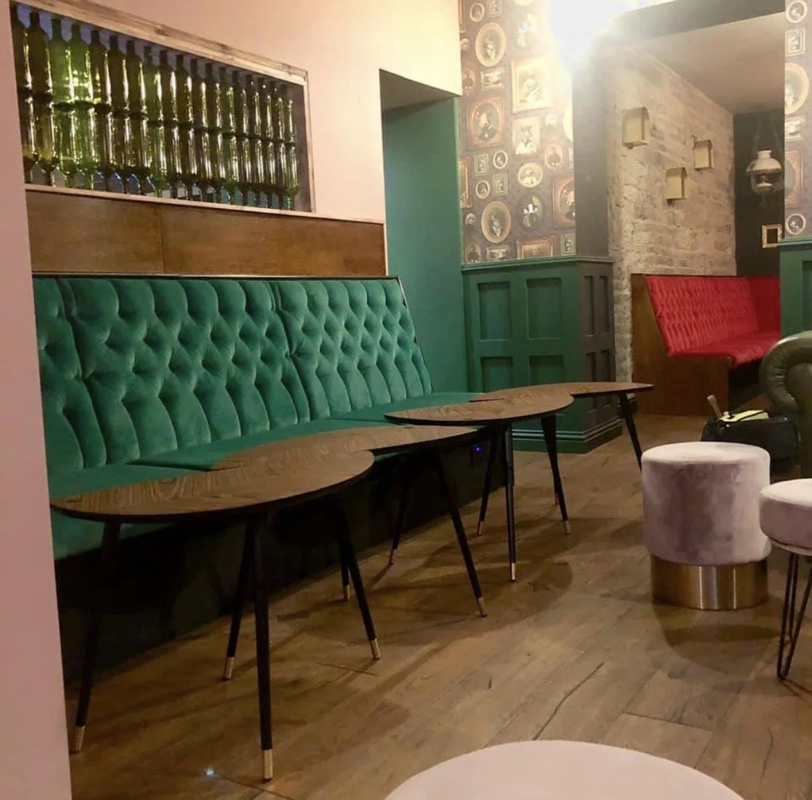 A cozy lounge with green tufted banquette seating, wooden tables, and pink and red velvet stools, featuring vintage wall decor and warm lighting.