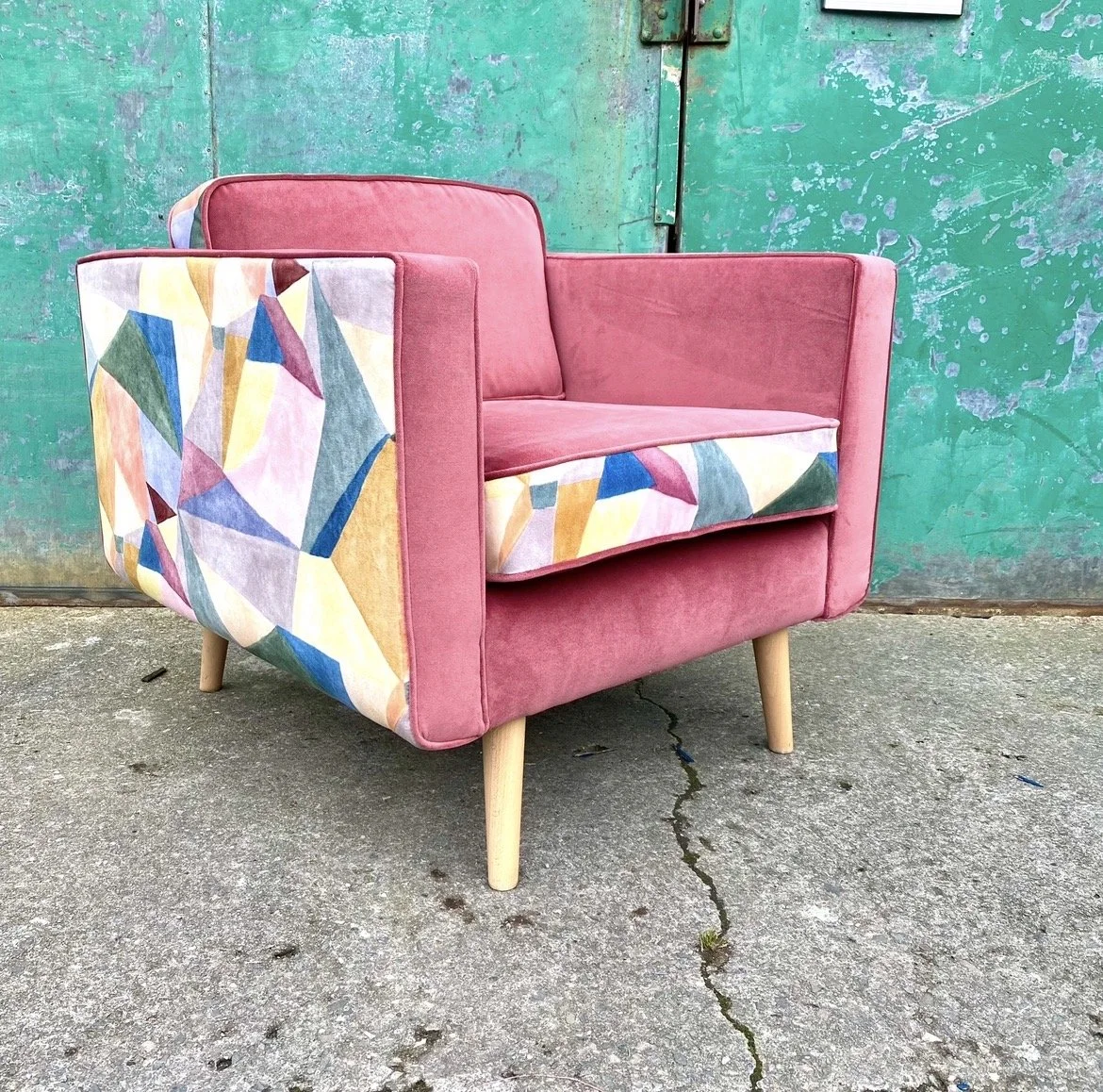 Pink armchair with patterned sides and backrest, wooden legs, placed on concrete ground in front of a green, weathered door.