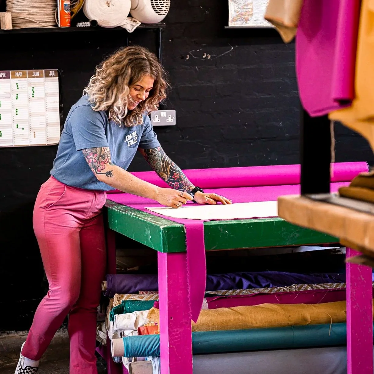 A woman with tattoos on her arms using a rotary cutter to cut fabric on a green and pink work table in a creative workspace.