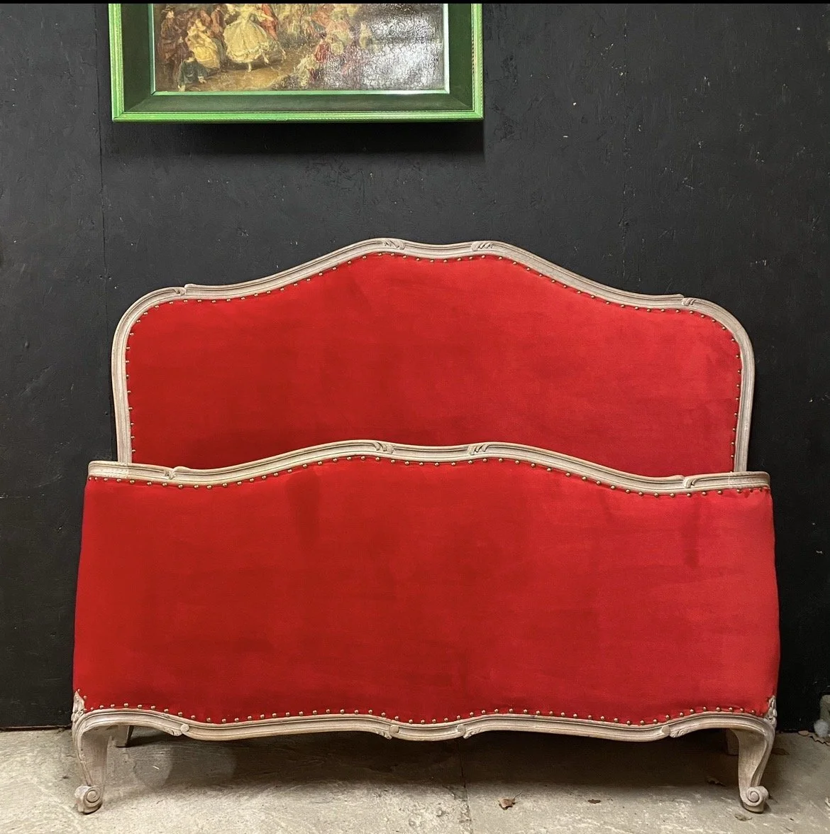 A vintage red velvet settee with a wooden frame against a black wall, with a framed painting above it.