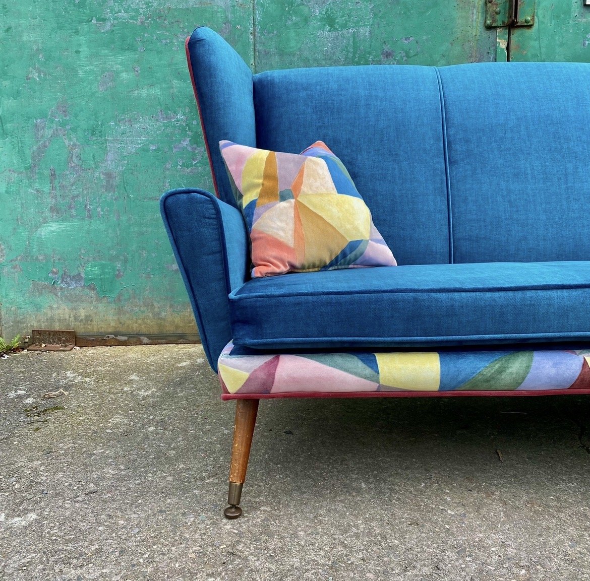 A bright blue vintage sofa with a colorful geometric-patterned pillow, outdoors on a concrete ground with a weathered green wall in the background.