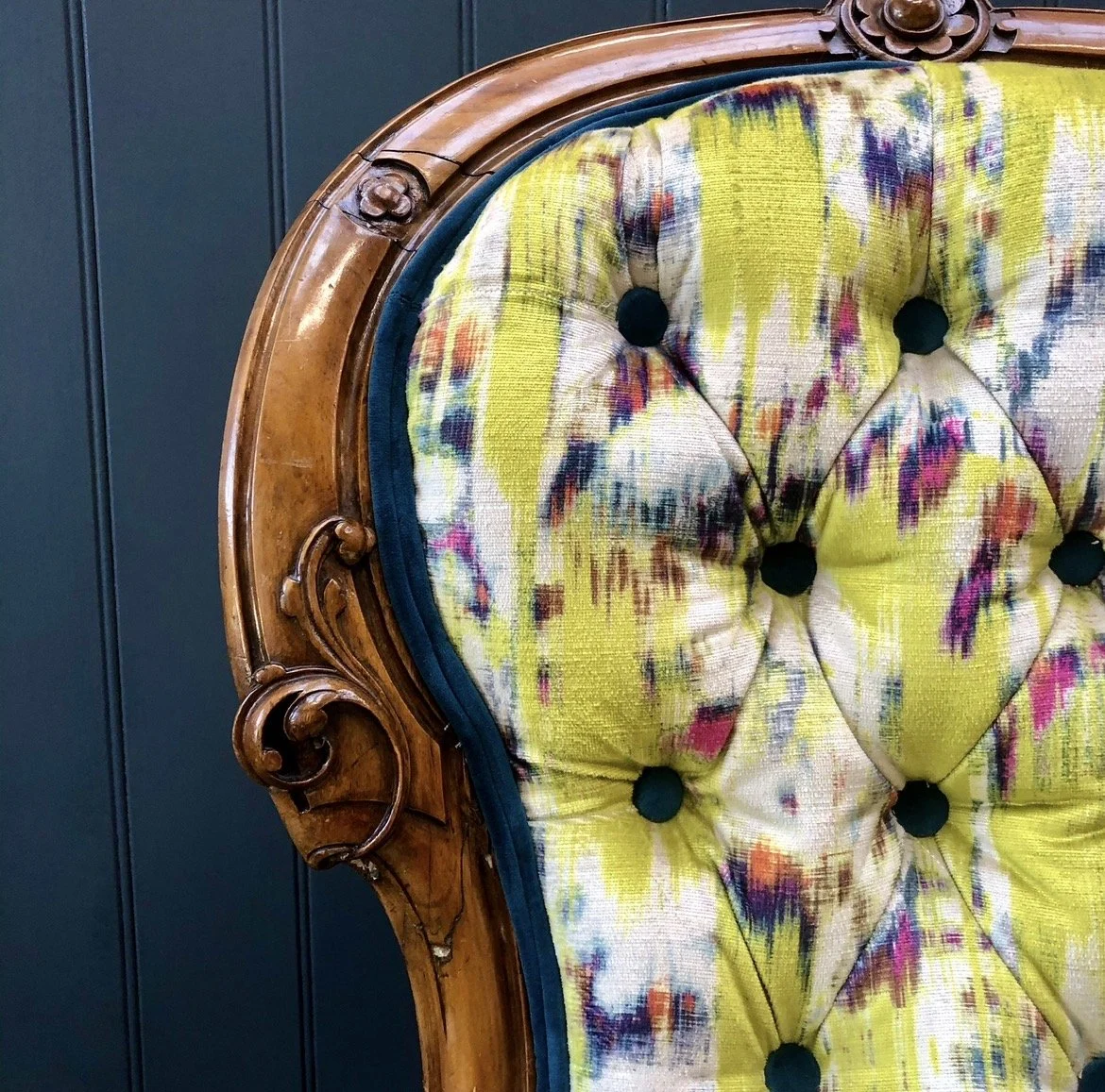 Close-up of a vintage wooden-framed chair with a colorful, tufted fabric upholstery in yellow, purple, and white patterns, with dark buttons.