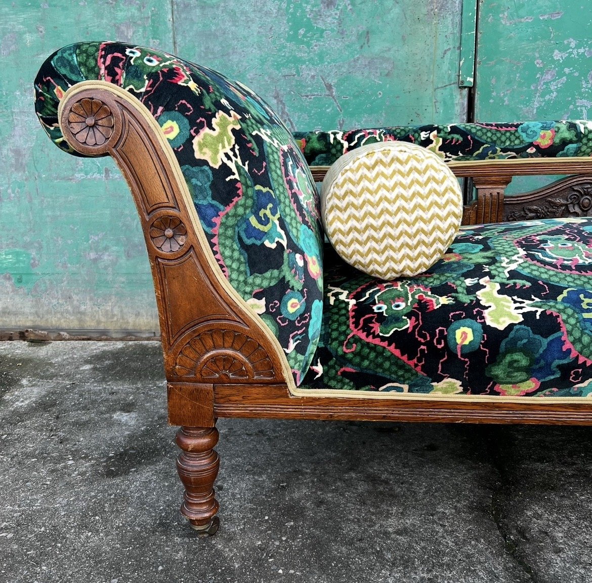 An antique wooden sofa with floral-patterned upholstery, a round cushion with a chevron pattern, and a green, textured backdrop.