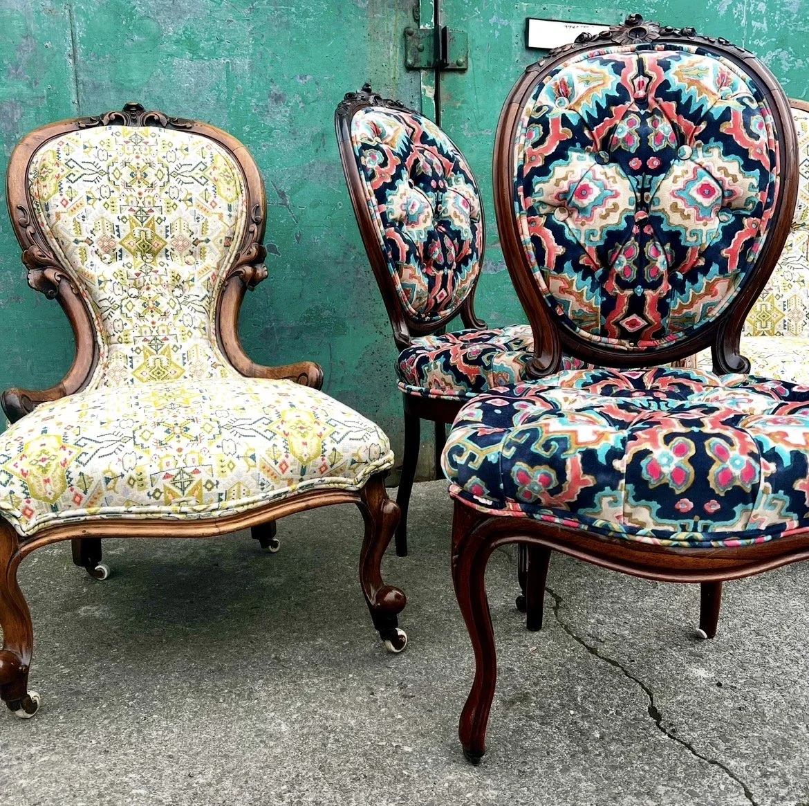 Three vintage upholstered chairs with ornate wooden frames, placed outdoors in front of a weathered green door.