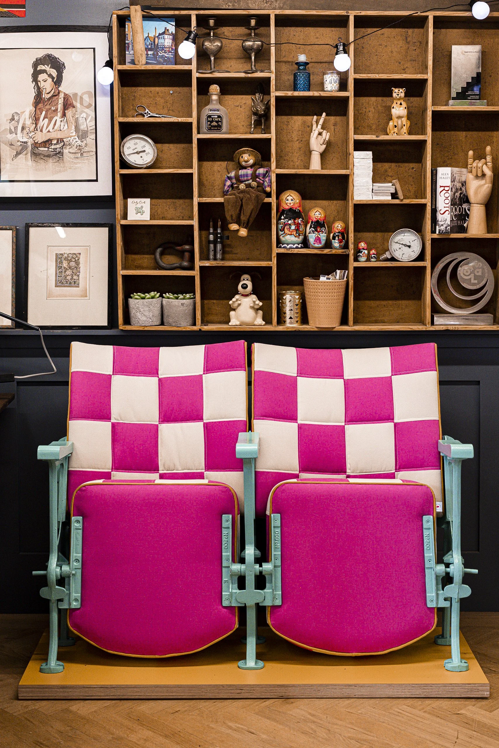 Two pink theater seats with light blue frames, placed on a wooden platform, facing away from the camera in front of a black wall with a wooden shelf filled with various decorative objects and artwork.